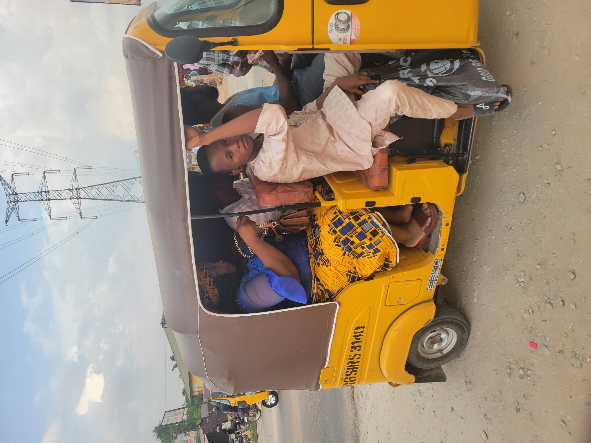The image shows a yellow "keke" or auto-rickshaw in Kwamba, Nigeria, packed with multiple passengers. One person is lying across the top of the seats, facing the camera. Another passenger is wearing a blue top and a yellow patterned skirt, holding a handbag. Other passengers are partially visible in the background. The rickshaw has "NG/SRS 3140" written on its side. Power lines are visible in the background, with buildings and vegetation along the roadside. The road surface appears unpaved.