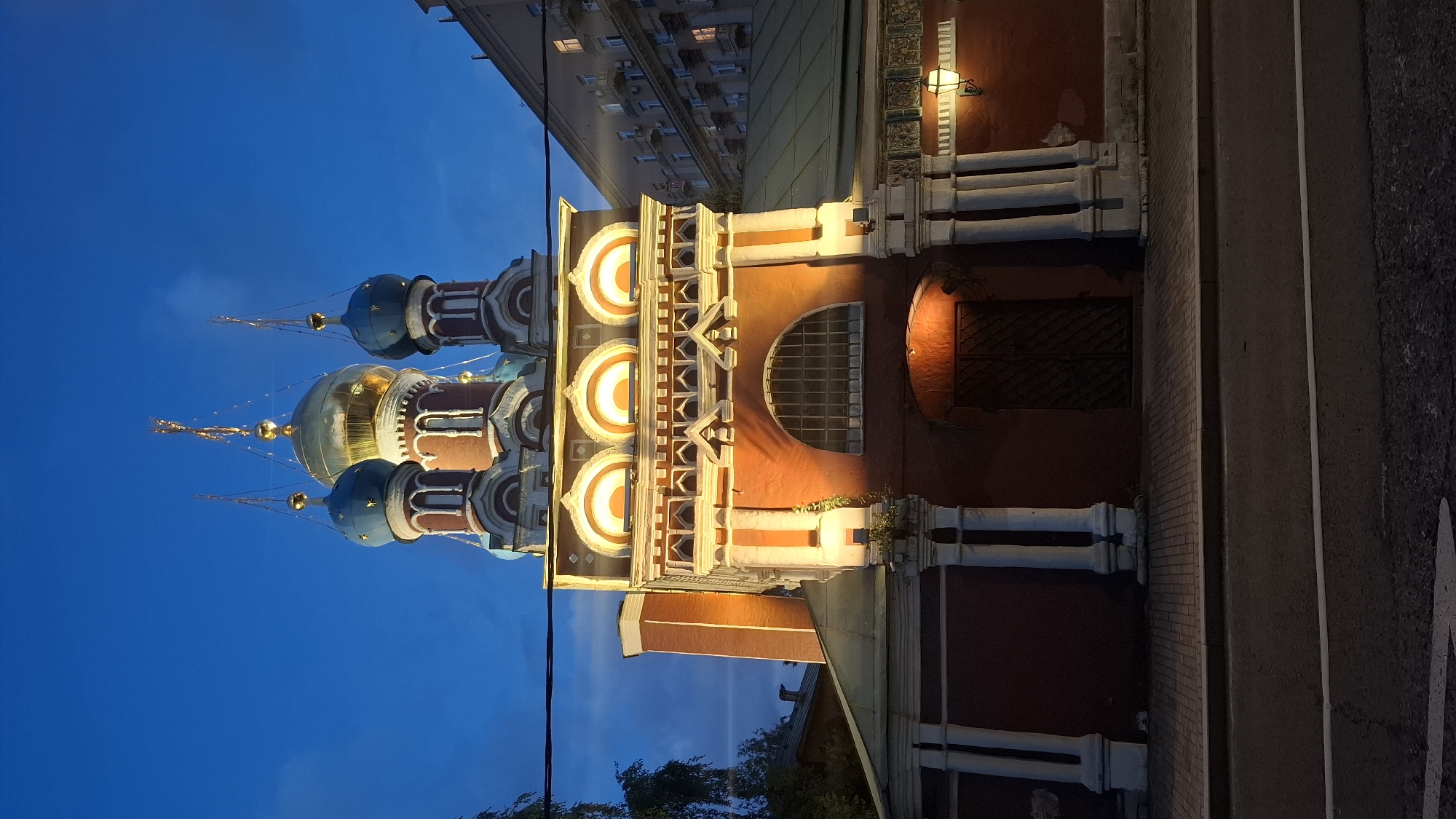 A Russian Orthodox church with multiple domes is lit up at dusk. The main dome is golden, and the smaller domes are blue. The facade of the church is red brick with white architectural details. There are arched windows illuminated by warm lights. A street with a sidewalk is in the foreground. An apartment building is visible to the right of the church. The sky is a deep blue with some wispy clouds.