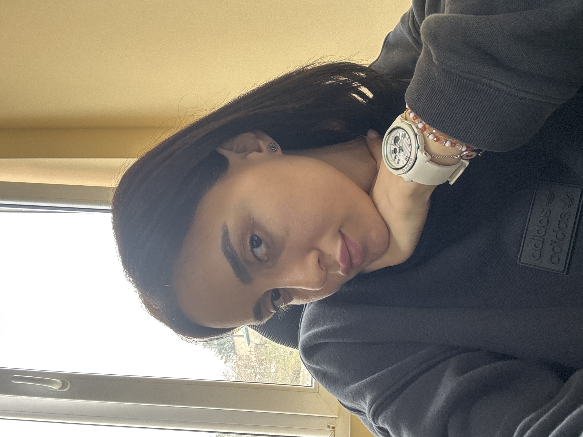 A female individual is depicted in a close-up, indoor setting within Tbilisi, Georgia. She possesses dark hair and dark eyes, looking directly at the camera with a neutral expression, her lips slightly parted. Her right hand is positioned to support her chin. She wears a dark gray or black crew-neck sweatshirt featuring two stacked black "adidas" logos in relief on the left chest area. On her left wrist, a white Casio Baby-G wristwatch with a white band is visible, along with multiple beaded bracelets in shades of red, white, and pink, and a small charm. A dark, circular stud earring is present in her right ear.
The background consists of a white uPVC window frame with a handle on the left, through which sparse trees and a distant building are visible under an overcast sky. To the right, a plain light yellow or beige wall is seen, along with the black mesh backrest of what appears to be an office chair behind the subject.