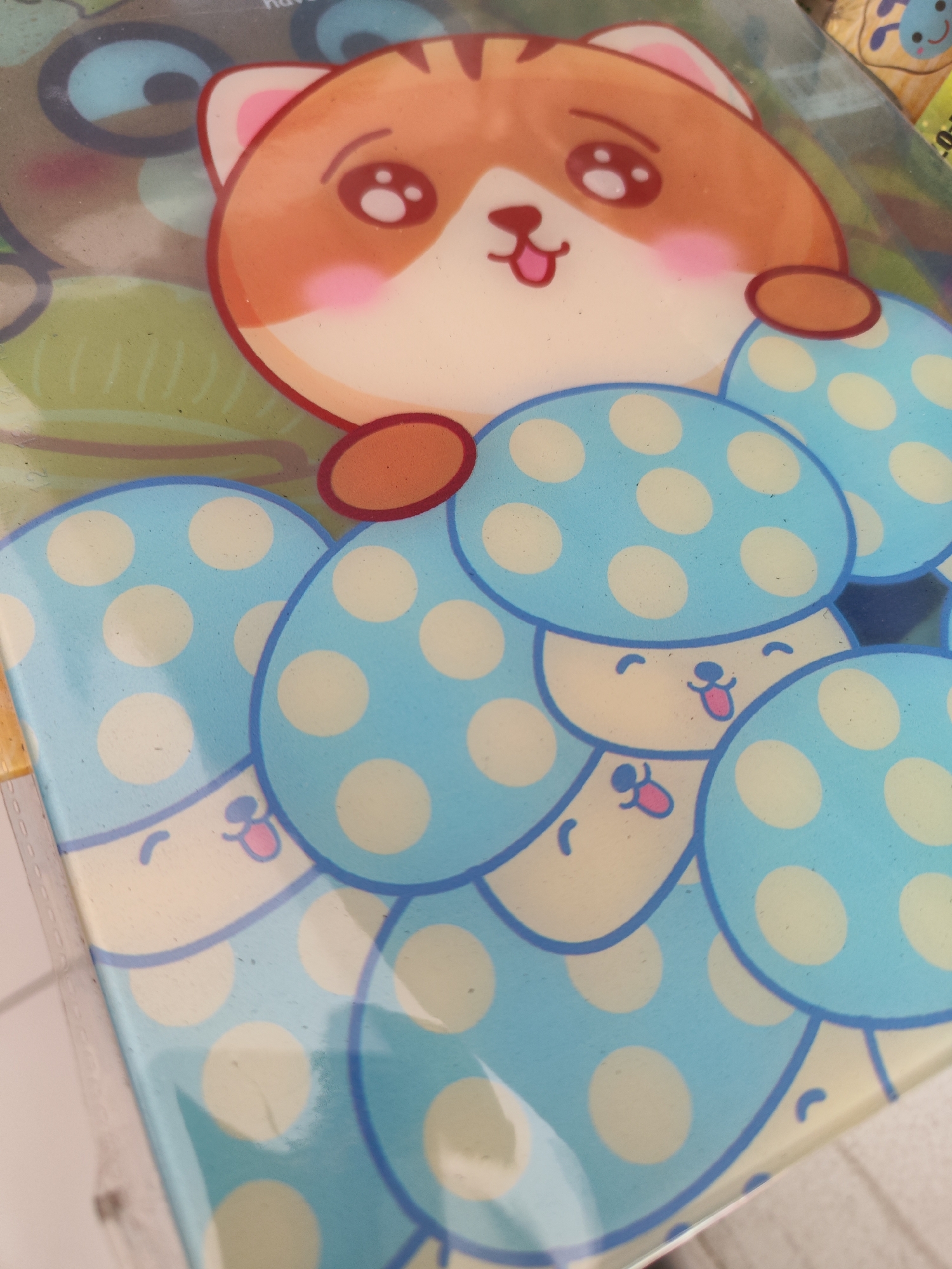 The image shows a close-up of a laminated notebook cover featuring cute cartoon characters. The main subject is a cat with orange fur and large, expressive eyes, peeking out from behind a cluster of blue mushroom characters. The mushrooms have yellow polka dots and smiling faces with tongues sticking out. The overall aesthetic is playful and childlike. The notebook cover appears to be new and is likely a stationery item. There is no visible indication of the city of Sragen, Indonesia, or any environmental context beyond the immediate object itself. The lighting suggests an indoor setting, possibly a store or a home.