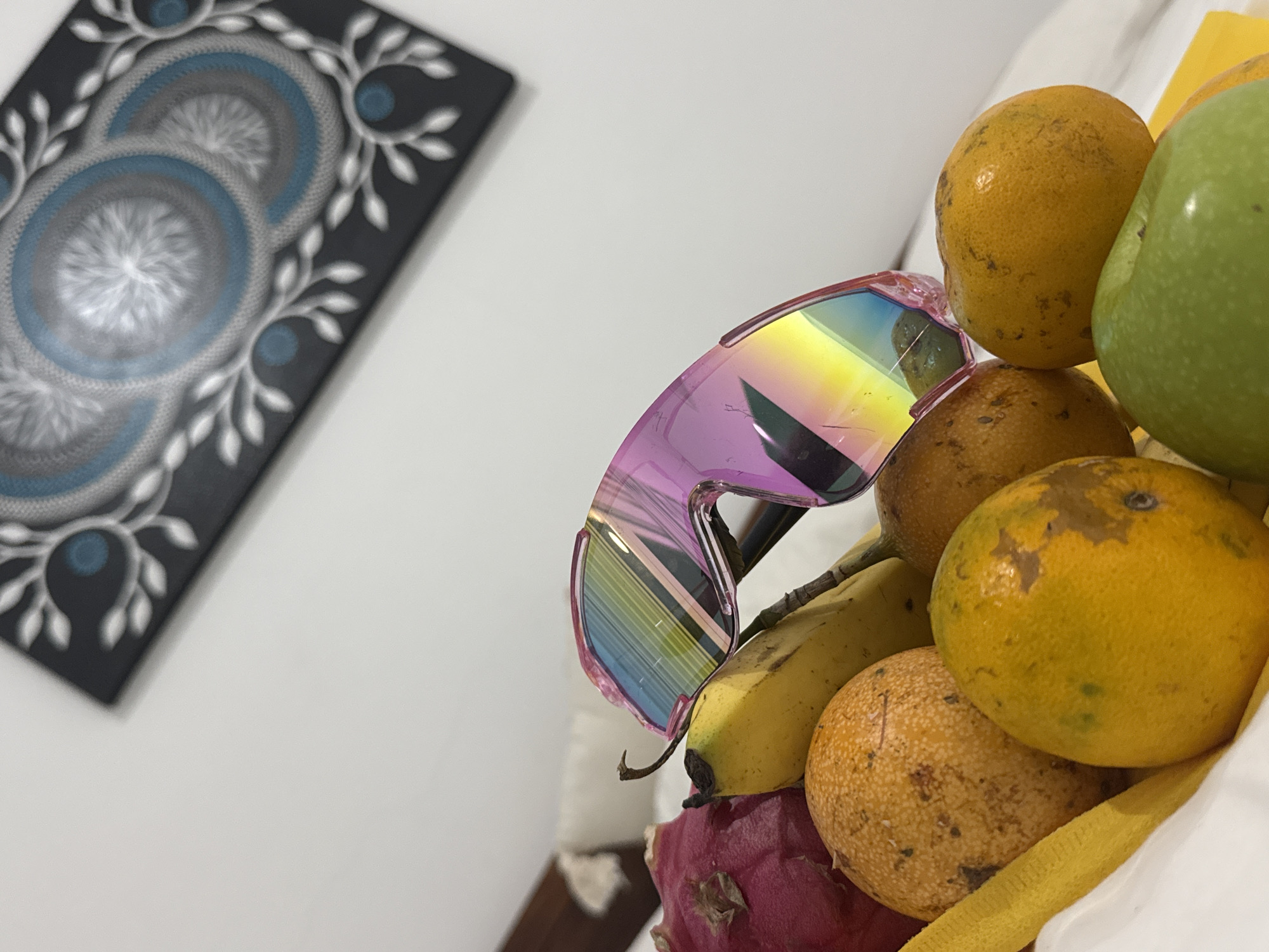 The image features a close-up of a collection of fruits and objects. A pair of pink and rainbow-colored sunglasses rests on a cluster of fruit, including oranges, passion fruit, a banana, a green apple, and a dragon fruit. The fruit appears to be piled together in a bag or container with a yellow cloth. In the background, a piece of wall art hangs on a white wall. The art features a black background with circular designs in shades of gray and blue, surrounded by white decorative motifs. The scene takes place indoors. There is no immediate evidence that this image was taken in Kuta, Indonesia.