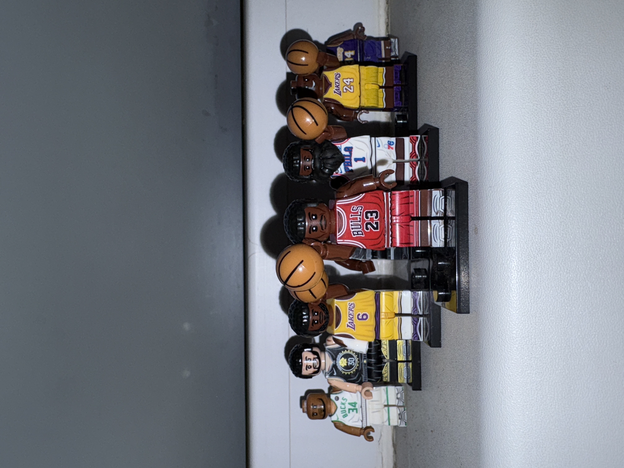 The image shows a row of Lego minifigures, each representing a famous basketball player. From top to bottom, they are wearing jerseys that represent: a Lakers player (#24), another Lakers player (#14), a 76ers player (#1), a Bulls player (#23), another Lakers player (#6), a Warriors player (#30), and a Bucks player (#34). Each is standing on a black Lego base and holding a brown Lego basketball, except the 76ers player, Warriors player, and Bucks player, who are not. The background is a combination of a gray and a white surface.
There is no visual information in the image to indicate its location is Izhevsk, Russia.