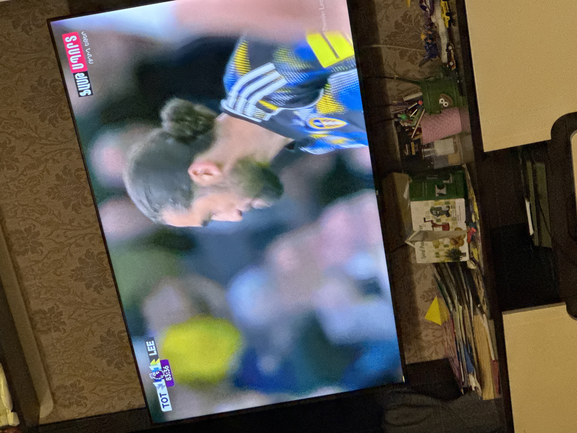 The image shows a television screen displaying a soccer game. The teams playing are Tottenham (TOT) and Leeds United (LEE), with the score being 1-1 at the 83:36 minute mark. The on-screen broadcast also features Armenian text ("Shant Premium"). The game appears to be part of the Premier League. The TV is placed near a wall with patterned wallpaper and a table with various items on it, including magazines, boxes, and toys. It is not possible to confirm the location as being in Yerevan, Armenia.