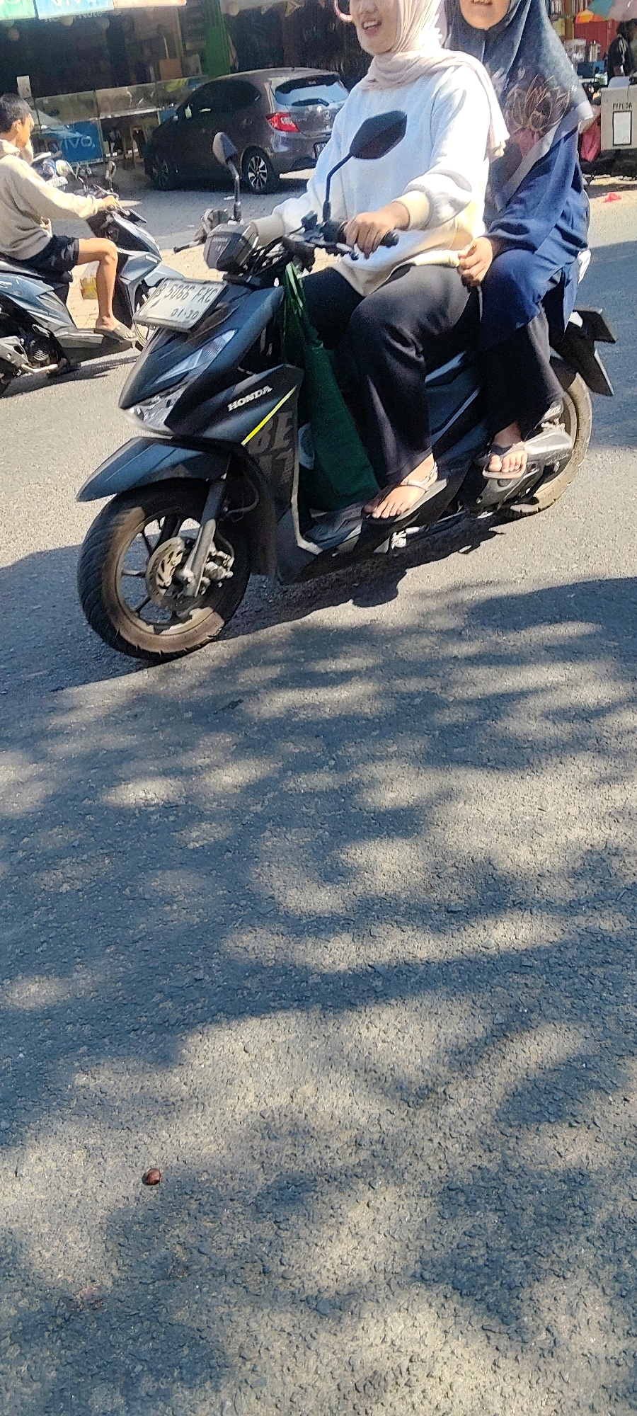 Two young women, wearing hijabs and casual attire, are riding a black Honda motorcycle on a paved road. The woman in front is wearing a white sweatshirt and dark pants, while the woman behind her is wearing a blue hijab and a navy blue garment. Both are wearing sandals. The motorcycle has a license plate with the characters "B 5066 FXC". In the background, a dark-colored car is parked, and other motorcycles are visible. The scene appears to be an urban environment, possibly a street in Cibatu, Indonesia, given the presence of common vehicles and casual pedestrian traffic. The road surface is asphalt, with shadows from overhead foliage cast across it. The lighting suggests it is daytime, with sunlight filtering through trees.
