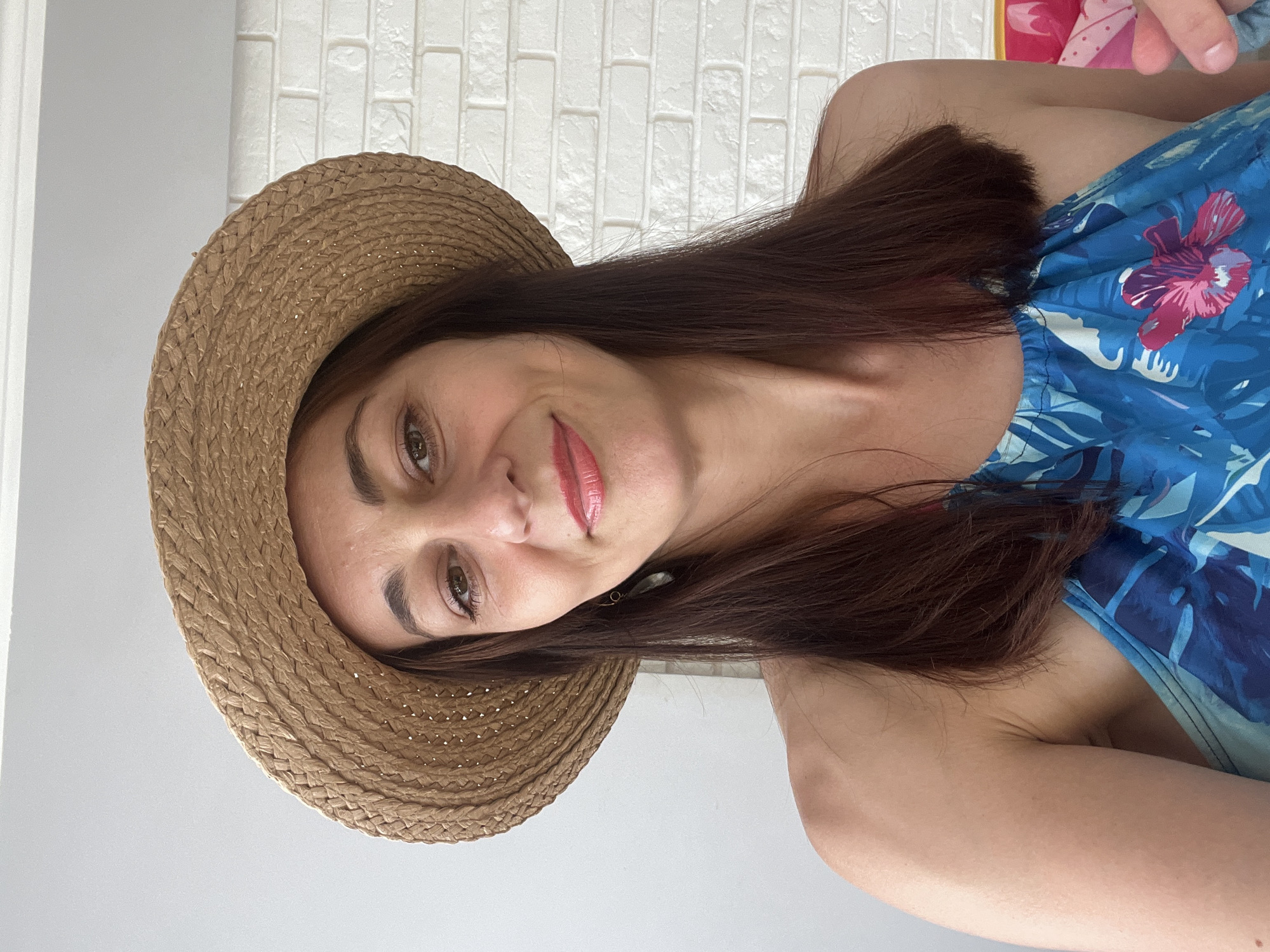 A woman wearing a straw hat and a blue floral print top is looking at the camera. She has long, dark brown hair and is wearing red lipstick. She is in front of a white brick wall.