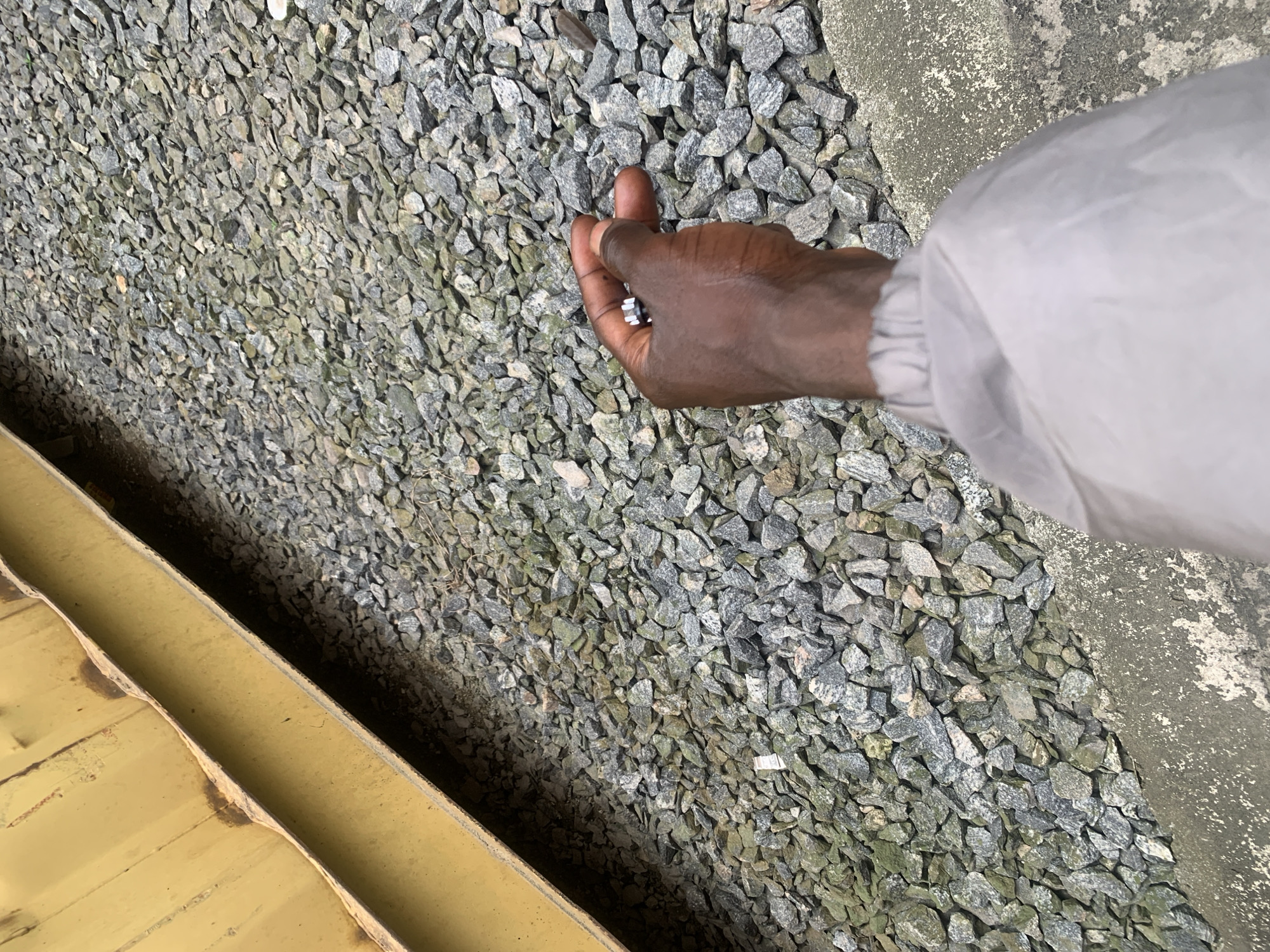 The image, set in Lekki, Nigeria, presents an overhead view of an outdoor ground area. The main subject is a person's dark-skinned hand, extending from a light gray sleeve on the right side of the frame. The hand is holding several small, silver-colored metallic objects, likely nuts or bolts, clustered in the palm and fingers.
The ground across most of the image is covered with a dense layer of irregularly shaped gravel or crushed stones, primarily in shades of gray with some greenish tints. To the far left, a section of a yellowish, corrugated metal structure, possibly a container or a part of an industrial building, runs parallel to the bottom-left edge. A narrow, dark strip separates this structure from the gravel. On the far right, a concrete surface or border, mottled with lighter patches, abuts the gravel.
The scene is illuminated by natural daylight, suggesting it is daytime. The presence of gravel, concrete, and the industrial-looking metal structure indicates an outdoor work environment, potentially a construction site or maintenance area. The person's hand holding fasteners suggests an activity related to assembly, repair, or handling of mechanical components. No text is visible in the image, and no specific emotions can be discerned.