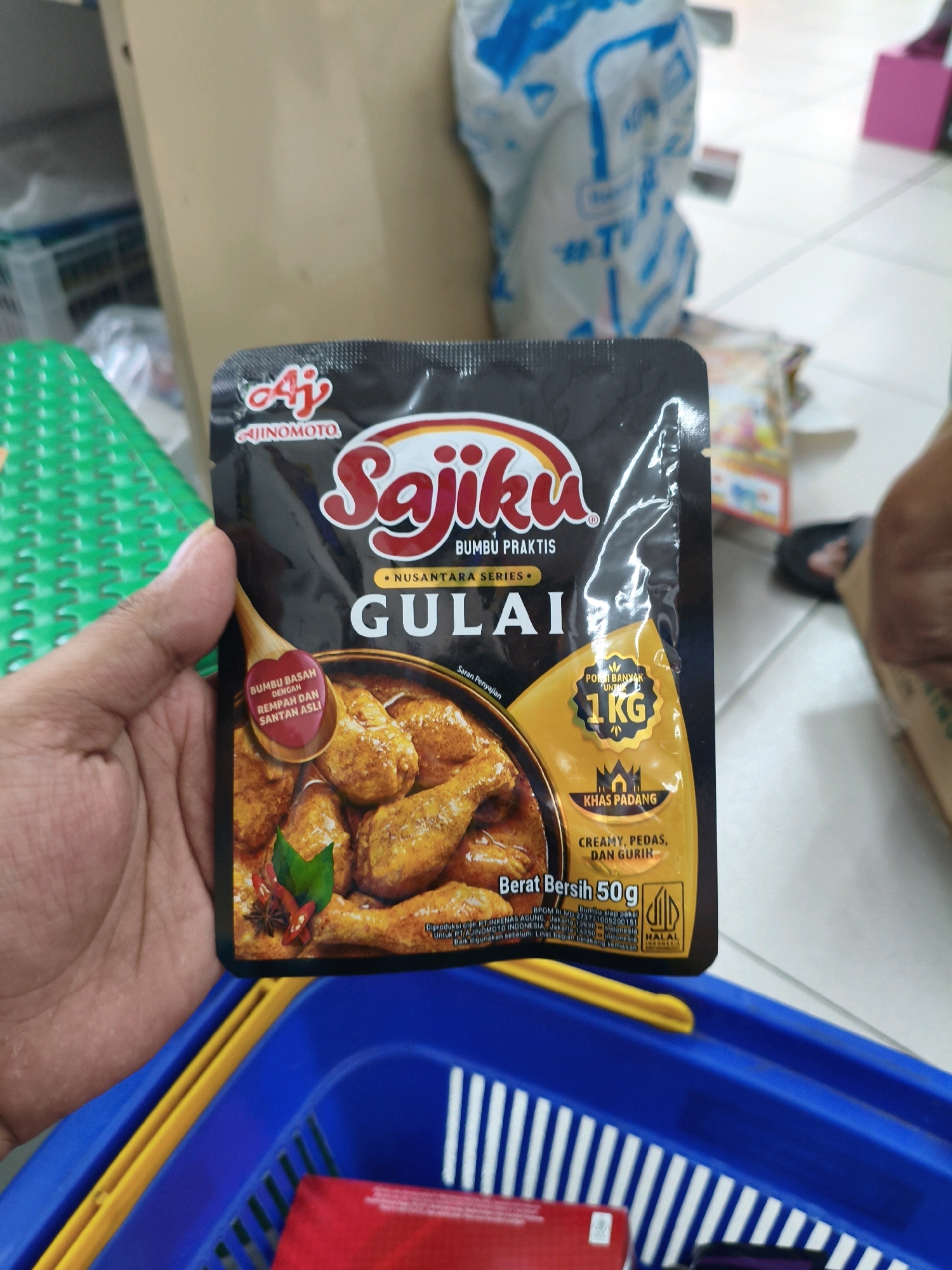 A hand holds a product package of Sajiku Gulai spice mix in the city of Sumberejo Kulon, Indonesia. The package features the Ajinomoto logo and an image of a rich, brown curry dish with chicken. Text on the package indicates it is a practical seasoning, suitable for 1 kg of meat, and is of the Padang variety, described as creamy, spicy, and savory. A red heart-shaped icon on a wooden spoon illustration on the package reads "Bumbu Basah dengan Rempah dan Santan Asli," meaning "Wet Seasoning with Original Spices and Coconut Milk." The product contains 50g of net weight and has a Halal certification. The background suggests an indoor retail environment, with a shopping basket visible in the foreground.