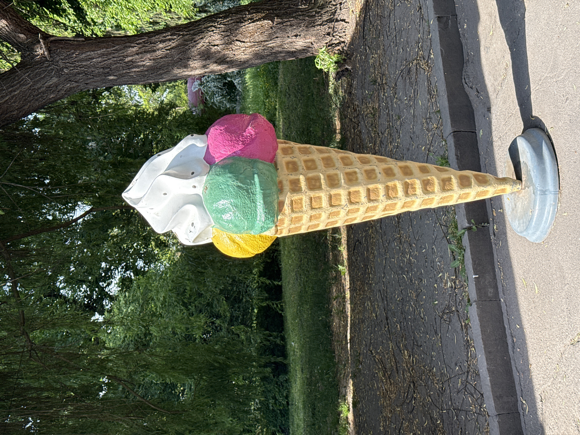 A large, oversized ice cream cone statue rests on a metal base adjacent to a sidewalk and a curb. The cone itself is yellow with a waffle pattern. It is topped with scoops of ice cream in white, green, pink, and yellow. The statue stands next to a tree and faces a grassy area. This image was taken in the city of Ternopil, Ukraine.