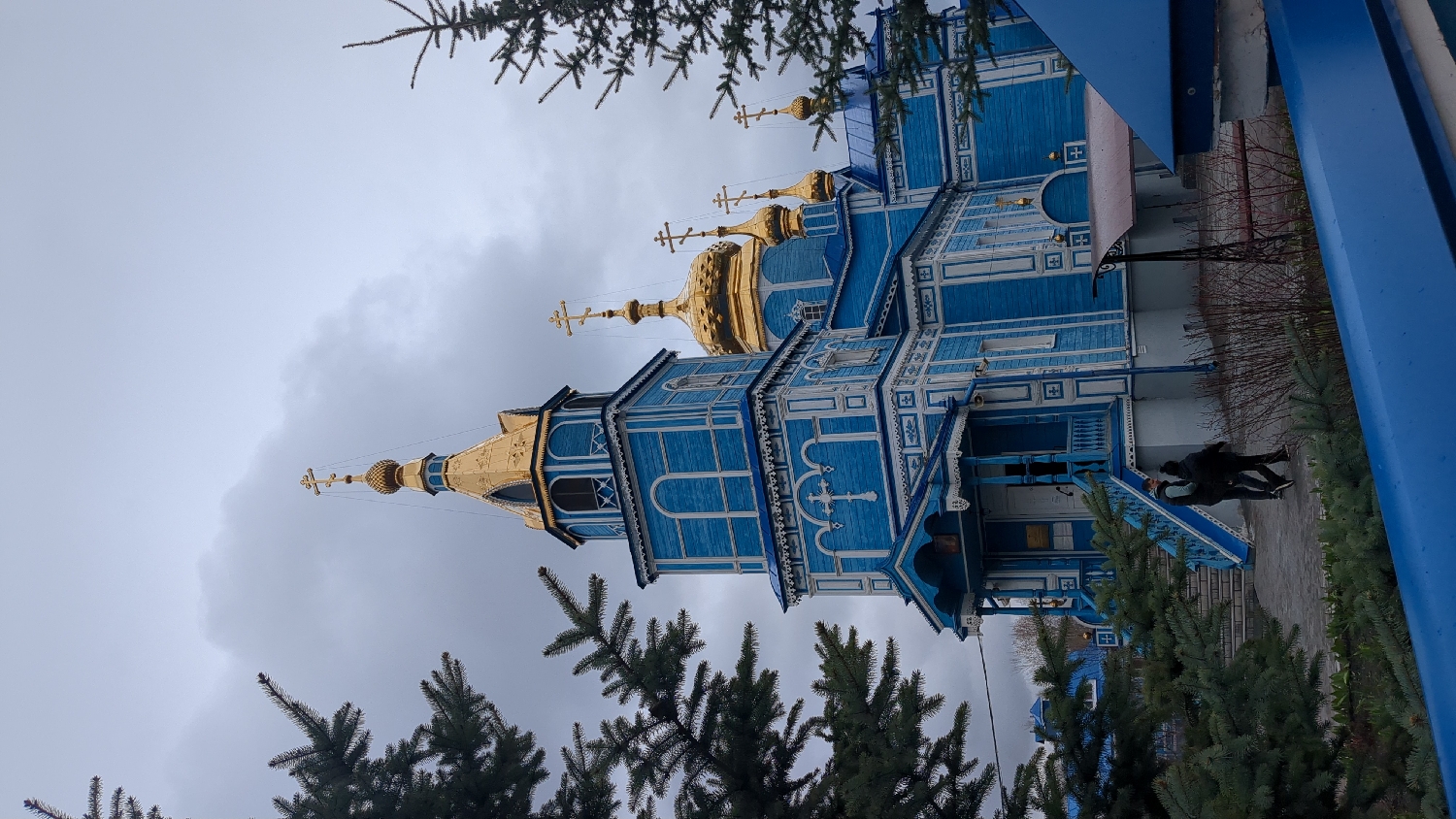 The image features a striking blue and white wooden Orthodox church under a gray, overcast sky. The church exhibits a classic Russian architectural style, characterized by multiple golden onion domes, each topped with a distinct Orthodox cross. Intricate white wooden carvings and decorative trim adorn the church's facade, windows, and cornices, creating a vivid contrast with the vibrant blue plank walls.
Two individuals, dressed in dark, casual clothing, are visible near the church's entrance on the lower right side. They stand on a paved area beside a set of blue wooden steps leading up to a covered porch. Green coniferous trees, likely fir or spruce, are prominent in the foreground and along the sides of the church, adding natural elements to the scene.
The cloudy sky suggests an overcast day, without direct sunlight. The detailed wooden craftsmanship and the distinctive domes are strong indicators of traditional Eastern Orthodox architecture, consistent with the location context of Ulyanovsk, Russia. There is no visible text in the image.