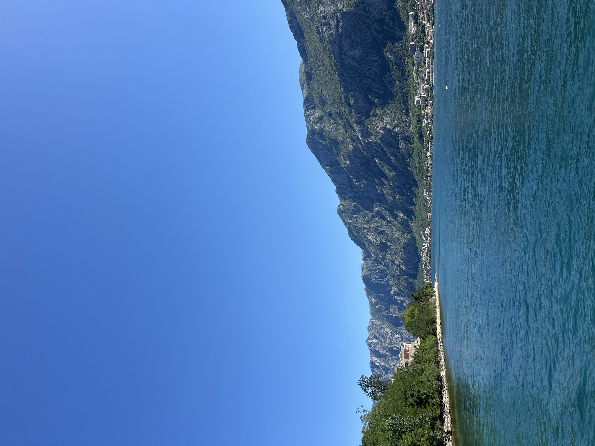 The image shows the landscape in Prčanj, Montenegro. On the right, there is a mountain range running along the coast, with a series of buildings and structures. The foreground features green vegetation and a shoreline. To the right of the shoreline is the body of water. To the left is a vast, clear blue sky.