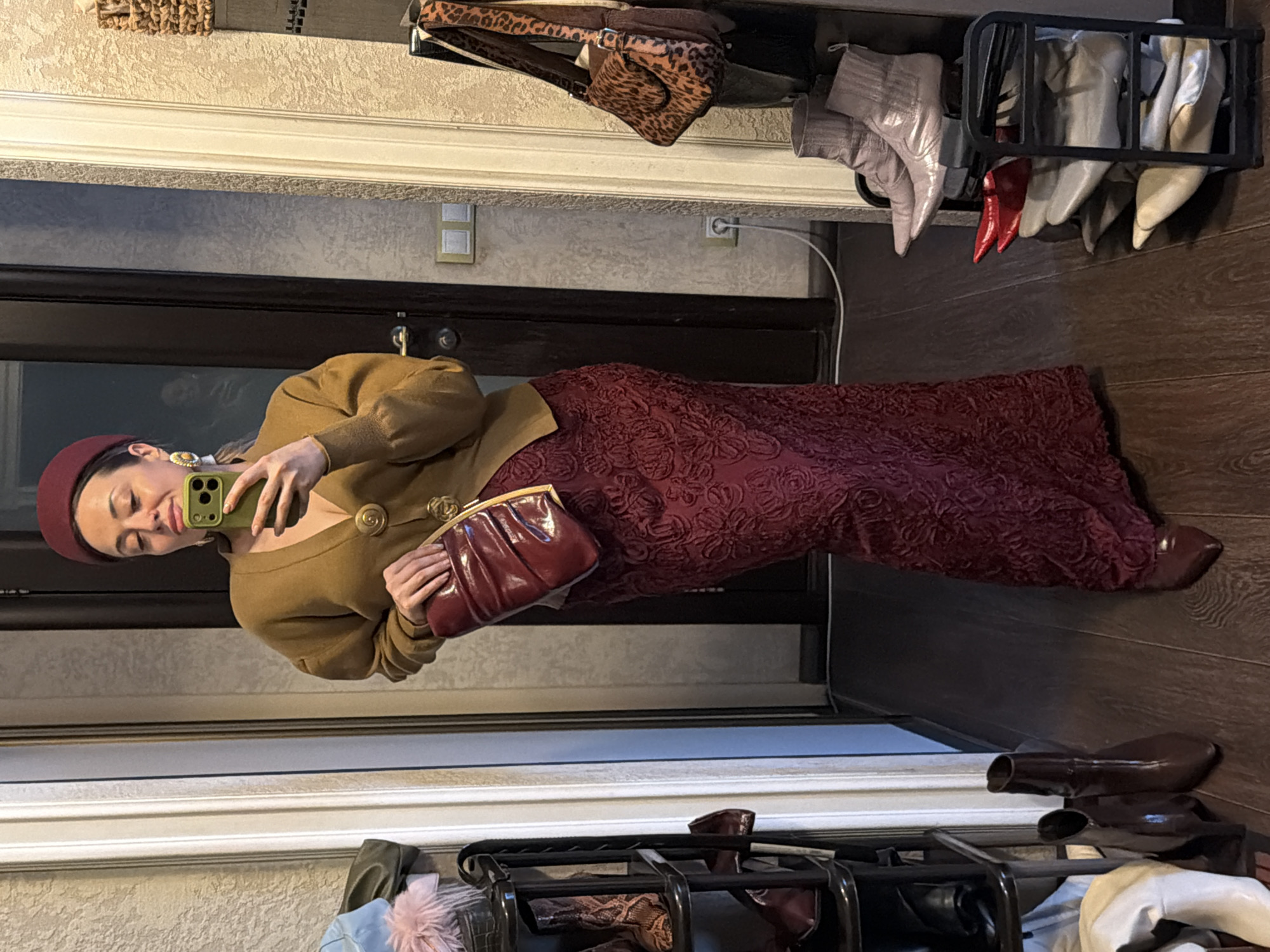 A woman in a maroon beret, mustard-colored cardigan, and maroon floral dress is taking a selfie in a mirror. She holds a green smartphone in her left hand and a maroon clutch in her right. The setting appears to be an entryway, with shoes organized in racks and on the floor. There is a door in the background. The location in the city of Krasnoyarsk, Russia is not identifiable from the visual elements in the image.