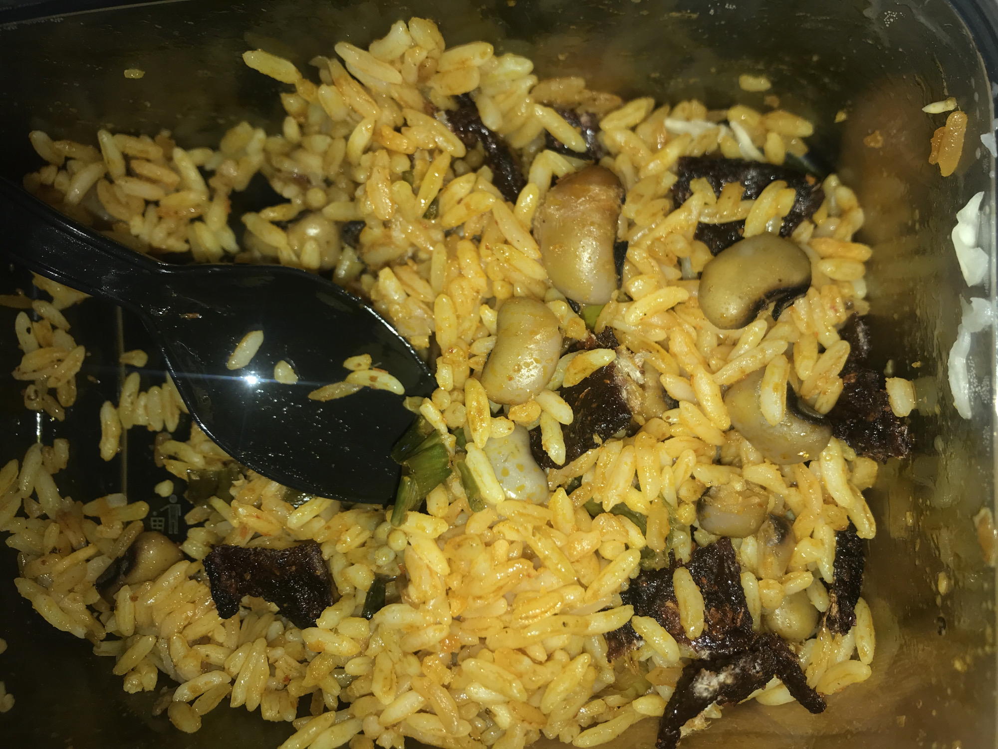 A close-up, top-down view captures a serving of a savory meal, likely in a dark, rectangular meal container. The main component is long-grain rice, which exhibits a warm yellowish-orange hue, suggesting it's seasoned or cooked with spices. Mixed throughout the rice are various other ingredients.
Several pale, oval-shaped beans, possibly white beans or light-colored legumes, are scattered within the dish. Interspersed with the rice and beans are numerous dark, irregularly shaped pieces. These appear to be chunks of a darker ingredient, potentially dried meat, mushrooms, or a deeply caramelized vegetable, adding a contrasting texture and color. Small green slivers, possibly chopped scallions or herbs, are also visible, contributing a touch of freshness.
A black plastic spoon is partially submerged into the food on the left side of the container, indicating the meal is ready for consumption or in the process of being eaten. The overall presentation suggests a hearty, flavorful dish. The lighting appears somewhat dim, creating subtle reflections on the container's surface. This type of rice and bean dish, potentially with added protein or vegetables, aligns with common meals found in regions like Rafin Par, Nigeria. There is no visible text in the image.