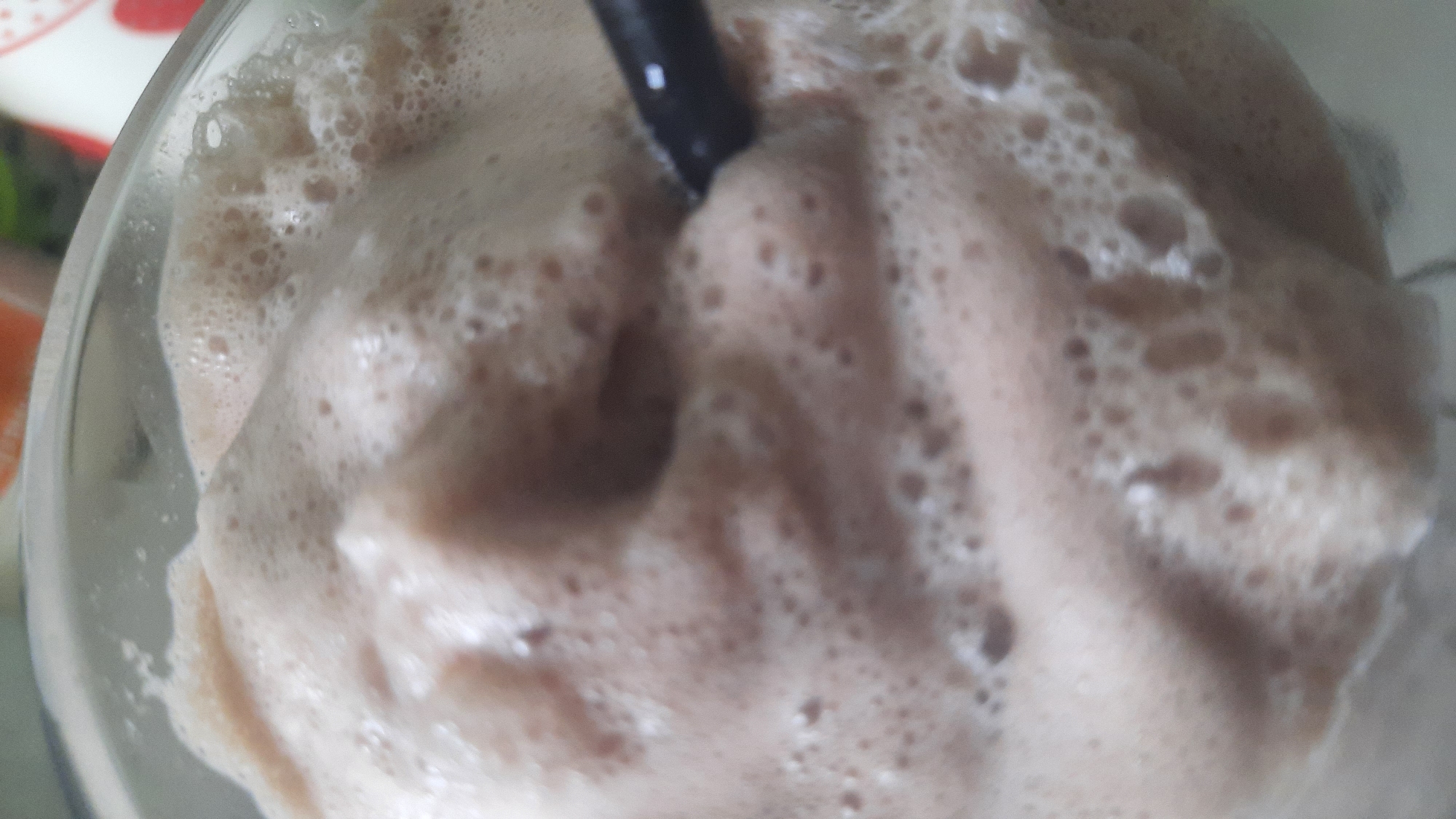 The image is a close-up, overhead view of a foamy, brown beverage in a clear glass. A black straw is visible on the right side, submerged in the drink. The beverage appears to be a milkshake or a similar frothy drink, possibly chocolate flavored, with numerous small bubbles on its surface. The lighting is soft, and the background is out of focus, with hints of red and green colors suggesting other objects or the environment. There is no indication of the location being Shklov, Belarus; the image depicts only the beverage and a straw.