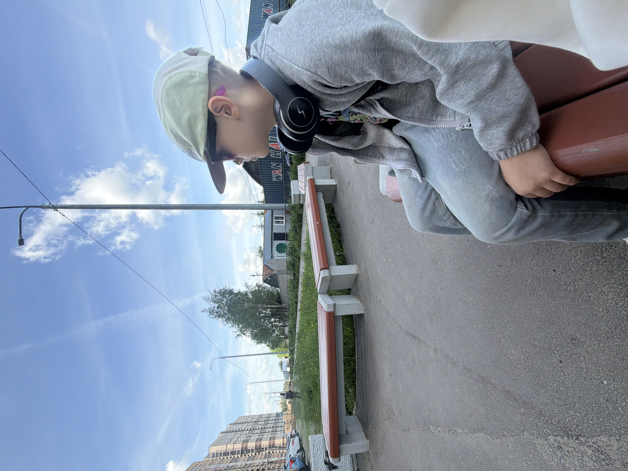 A young person, likely a child or pre-teen, is shown sitting on a reddish-brown and concrete bench in an urban or suburban outdoor setting. They are viewed from the side, wearing a light-colored baseball cap, a grey zipped hoodie, and light blue jeans. Black over-ear headphones are around their neck, and a small pink device (possibly a hearing aid or earbud) is visible in their right ear. The subject is looking downwards, with their right hand resting on the bench seat.
The scene is brightly lit under a clear blue sky with scattered white clouds, indicating daytime. A paved path extends in front of and alongside the bench, with a strip of green grass and several other identical benches visible further down. In the background, there's a tall, multi-story building (likely residential) on the left. To the right, a dark, container-like structure is visible, bearing the white text "WAN HAI". Trees, bushes, utility poles, and overhead wires complete the background. The overall impression is a quiet moment in an everyday city environment, consistent with a location such as Saint Petersburg, Russia.