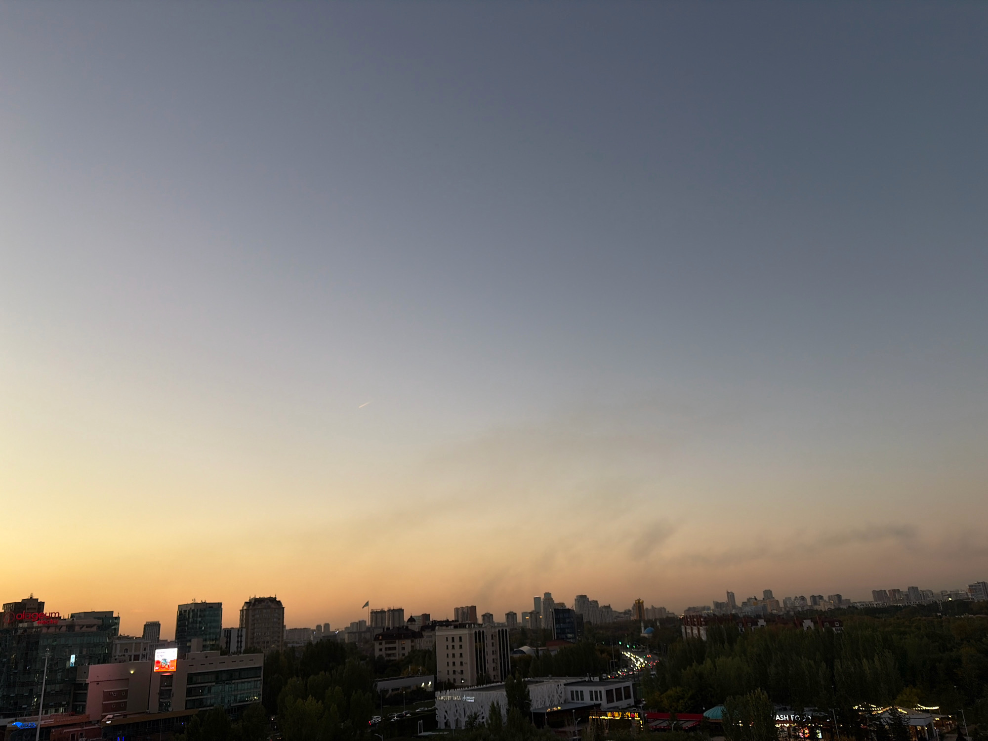 This is an eye-level, wide shot of a city skyline at dusk, likely Astana, Kazakhstan. The sky transitions from a pale yellow at the horizon to a light gray at the top. A faint contrail is visible in the upper left portion of the sky. Below the sky is a dense collection of buildings of varying heights and architectural styles, with trees in the foreground. On the left side of the image, a modern building with glass windows and illuminated signage is prominent. Further to the right, other buildings, some appearing residential and others commercial, are visible. Traffic lights and streetlights suggest ongoing activity in the city. A flag is visible on a pole atop one of the buildings. The overall impression is one of an urban environment at the end of the day.