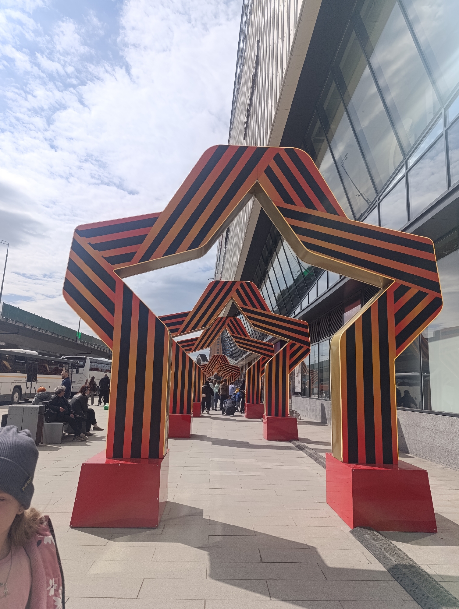 The image shows a pedestrian walkway in Moscow, Russia, lined with five-pointed star-shaped arches in a red and black striped pattern. The arches have gold trim and rest on red bases. People are walking under the arches, and buses and people are visible in the background. A modern building with reflective windows is on the right side of the walkway. People are sitting on benches. The sky is partly cloudy.