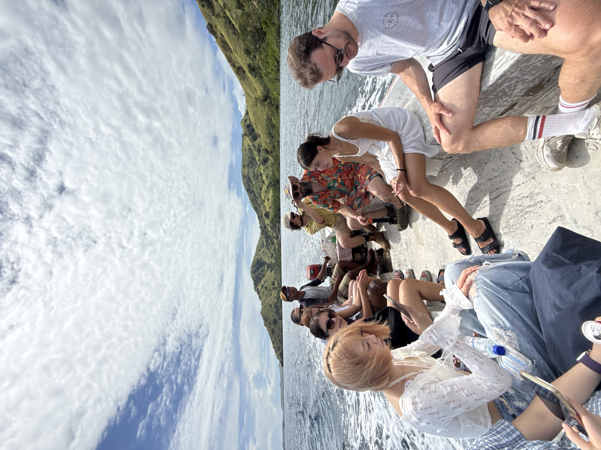 The image shows a group of approximately 12 people seated on a boat, seemingly on the water off the coast of a mountainous island. The sky above is filled with clouds. The boat is moving, as indicated by the wake in the water. People are mostly dressed in casual clothing. Some are looking at their phones, some are looking at the water. The location is specified as Unknown, Indonesia.
