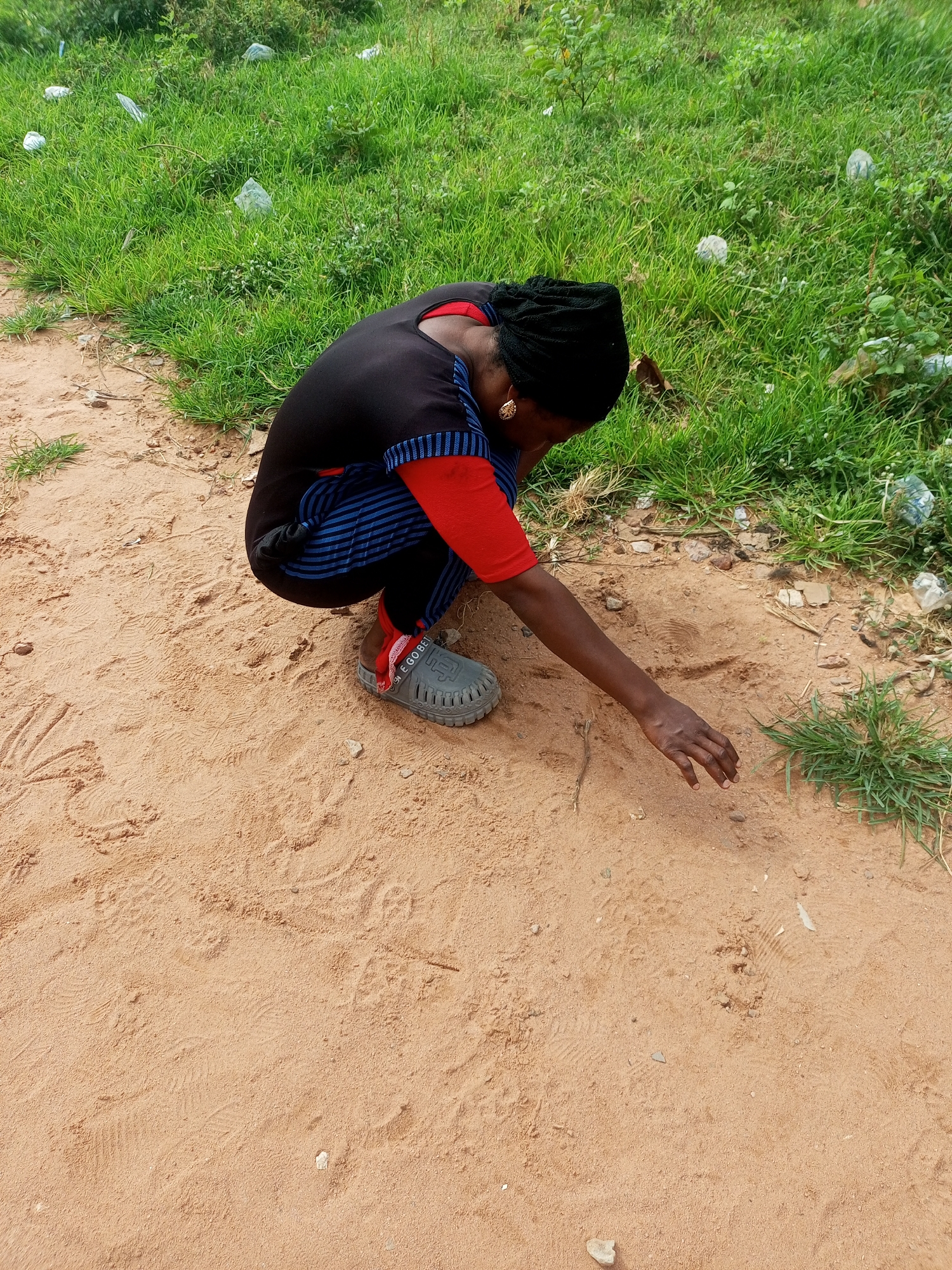 In the city of Tempure, Nigeria, a person is seen squatting on a sandy path next to a grassy area. The individual is wearing a black headscarf, a black shirt with blue stripes and red sleeves, and grey slip-on shoes with the inscription "E GO BELIEVE". The person is reaching down towards something on the sandy ground. The grassy area appears somewhat unkempt and contains scattered pieces of white plastic debris. The sandy path shows faint impressions, possibly from footprints. The overall scene suggests an outdoor environment with signs of littering.