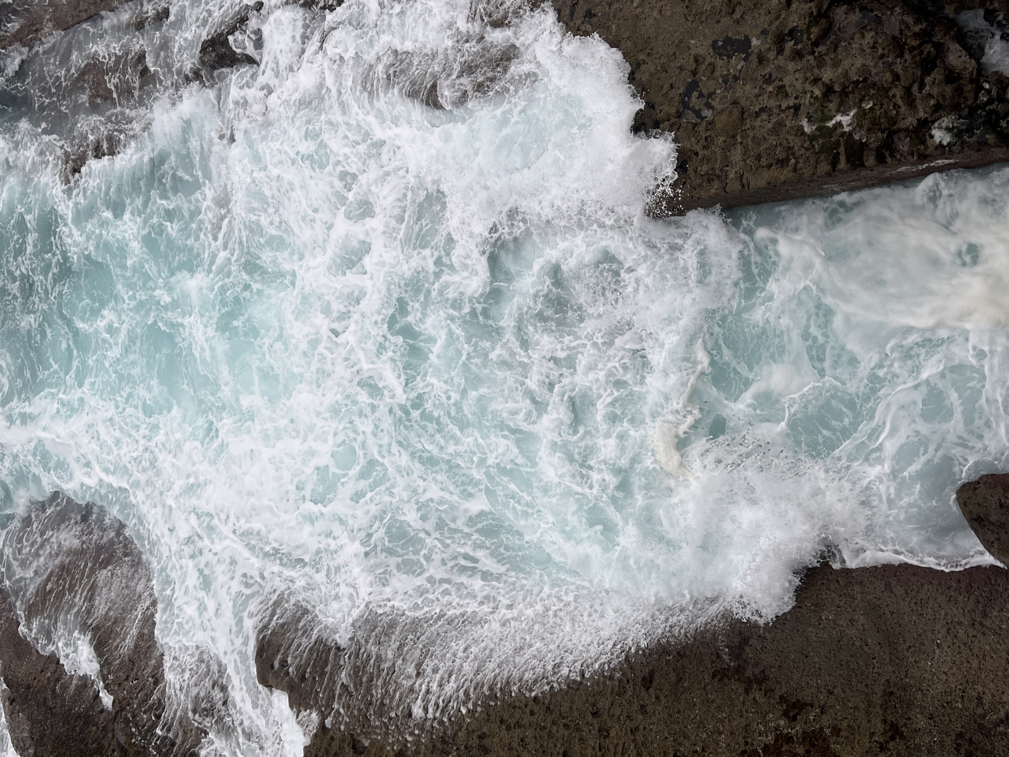 The image is an aerial, close-up view of ocean water churning against a rocky shoreline. The water is a vibrant turquoise with white foam and waves crashing against the dark, textured rocks. The rocks appear jagged and wet, with some areas showing darker, possibly submerged portions and lighter, exposed surfaces. The waves are dynamic, creating a sense of movement and power in the scene. There are no people or man-made objects visible. The environment is natural, characterized by the interaction of water and rock formations. Based on the visual characteristics, this scene is consistent with the rocky coastline found in areas such as Monte Urgull in San Sebastián, Spain, where the Atlantic Ocean meets the rugged land.