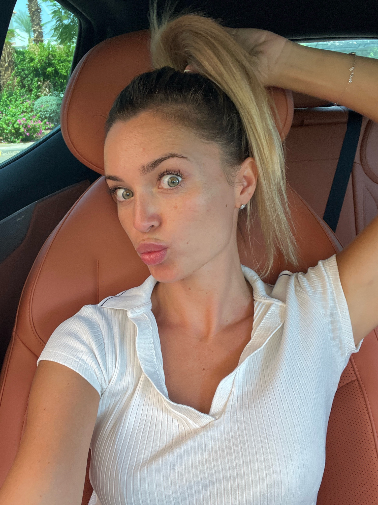 A young Caucasian woman with blonde hair styled in a high ponytail is sitting in the driver's seat of a car. She is looking directly at the camera with wide, green eyes and puckered lips, making a kissing gesture. Her right hand is raised, holding a portion of her ponytail. She is wearing a white, ribbed, short-sleeved polo shirt and a delicate silver bracelet on her left wrist. The interior of the car features brown leather seats. Outside the car window, lush green foliage and palm trees are visible, suggesting a warm, sunny climate. The scene is consistent with a daytime photograph taken in a car in Sharm El Sheikh, Egypt, given the presence of palm trees and the characteristic sunny weather.