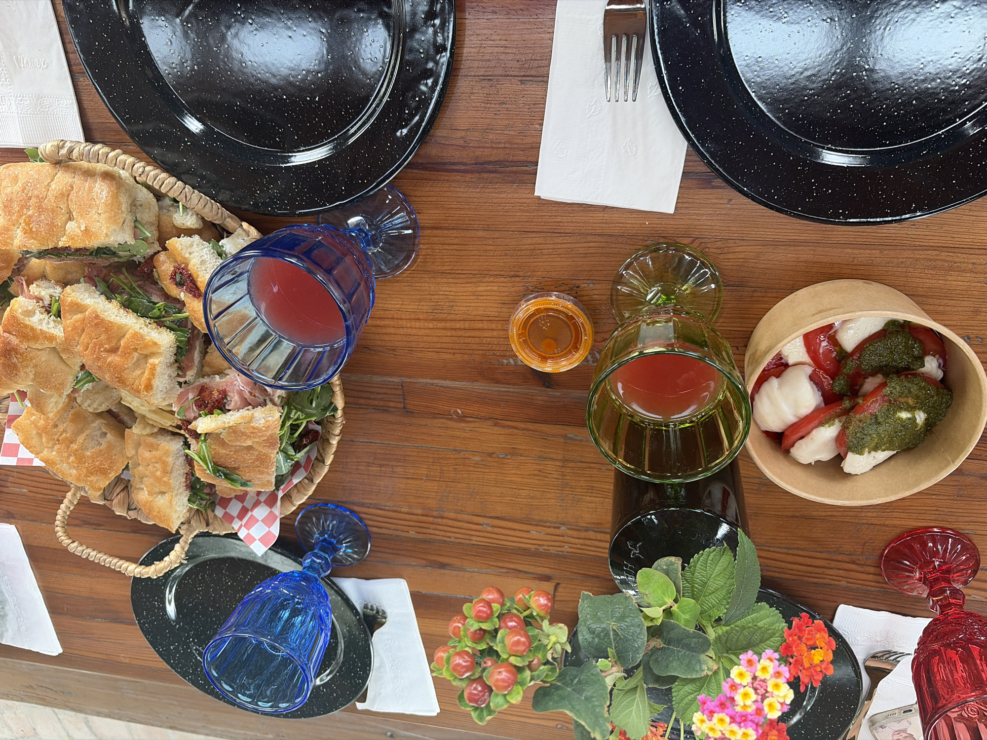 A wooden table is set with various items for a meal. Two black speckled plates sit near the top corners, one with a fork and a white napkin. A basket overflowing with sandwiches is in the upper left corner. Three glasses, each of a different color (blue, green, and red), are placed around the table, two of them lying on their sides. A small container of what appears to be dip is near the center of the table. There is a small bowl containing mozzarella and tomatoes topped with pesto. A cluster of small red flowers and other greenery decorates the lower center of the table.