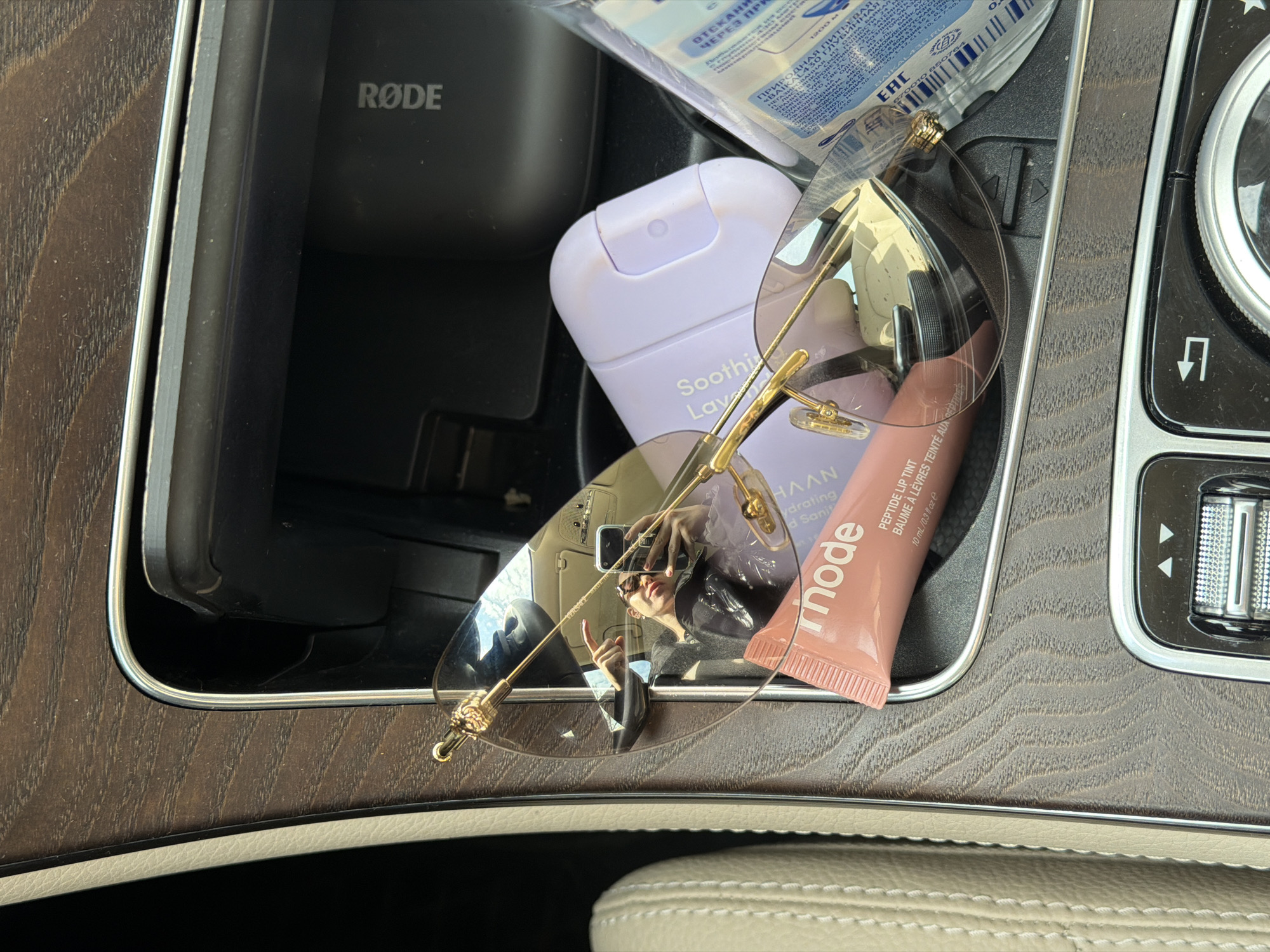 The image shows the inside of a car console, likely a luxury vehicle given the interior finishes. A Rode microphone holder is visible in a compartment. Lying on top of the console are several personal care items: a bottle of Haan "Soothing Lavender" hydrating hand sanitizer, a "rhode" Peptide Lip Tint, a clear package with what appears to be a bottled water of the brand "Baikal 430" with text in Russian, and a pair of sunglasses with gold frames. The sunglasses reflect a person wearing a black jacket and sunglasses, holding up a hand. The environment consists of the car's interior, with faux-wood trim and light-colored leather seating. The location cannot be determined with certainty to be Moscow, Russia, based on the contents of the image alone, but the presence of the "Baikal 430" bottled water, with text in Russian, suggest that location.