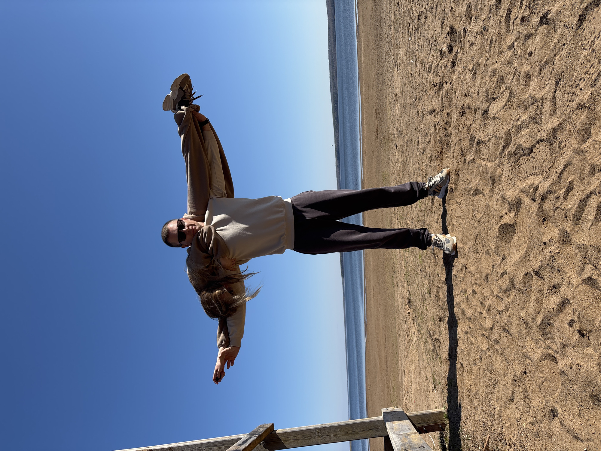 A person in a light beige sweatshirt and dark pants is striking a pose on a beach in Chaikovsky, Russia. They are upside down, with their head pointing towards the clear blue sky and their feet on the sandy beach. The sea is visible in the background. The person is wearing sunglasses and appears to be holding a brown jacket or coat above their head. They are positioned near a wooden structure. The beach is sandy with visible footprints.