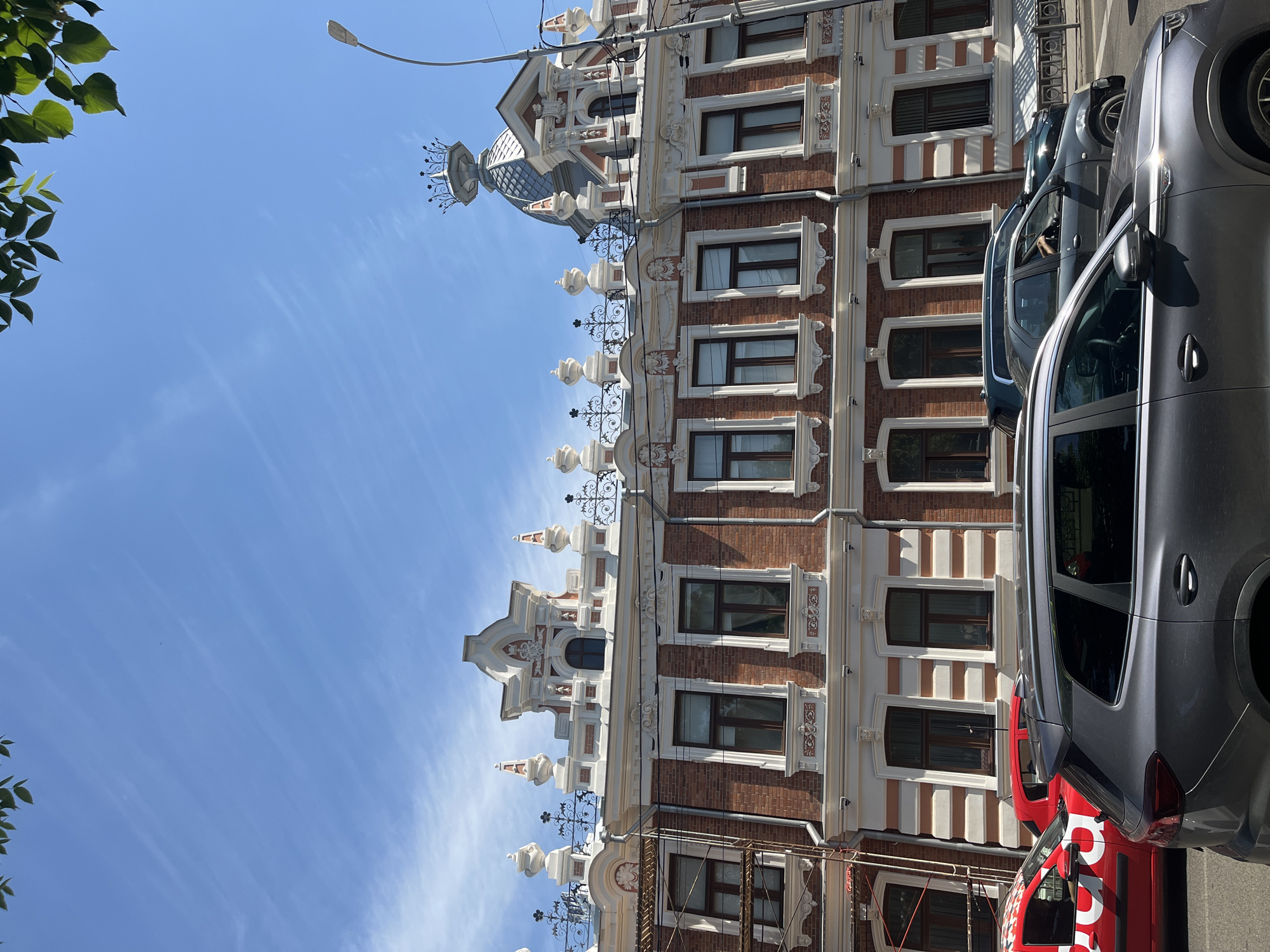 The image depicts the exterior of a historic brick building in Krasnodar, Russia, with ornate white stucco detailing and a domed roof topped with a crown-like structure. Several cars are visible on the street in front of the building, including a red car with a logo and a grey SUV. The sky is blue with some wispy clouds, and green leaves from a tree overhang the top of the frame. The building appears to be a multi-story structure with numerous windows. The overall scene suggests a sunny day in an urban environment.