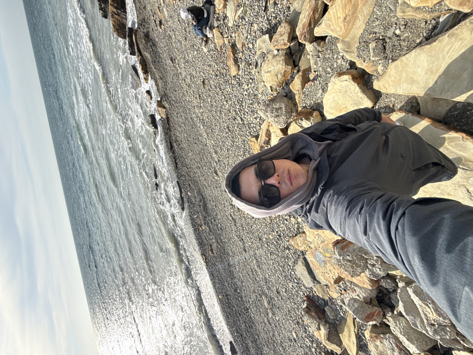 The image depicts a coastal scene in Anapa, Russia. In the foreground, a male individual is taking a selfie, holding the camera at arm's length. He wears dark sunglasses, a grey hooded sweatshirt with the hood up, and a dark grey jacket. He stands among large, light-colored rocks and boulders. In the midground, further down the beach towards the waterline, a second individual is seated on the pebble beach, facing away from the camera. This person wears a white cap or hood and a dark jacket, sitting on what appears to be a dark mat or bag. The beach consists of numerous small to medium-sized grey and brown pebbles, with larger rocks intermittently along the shoreline. The sea occupies the upper half of the frame, exhibiting choppy waves and a bright reflection of sunlight across its surface, particularly on the left side. The sky above the horizon is visible, appearing clear or lightly hazy. The overall environment is illuminated by bright daylight.