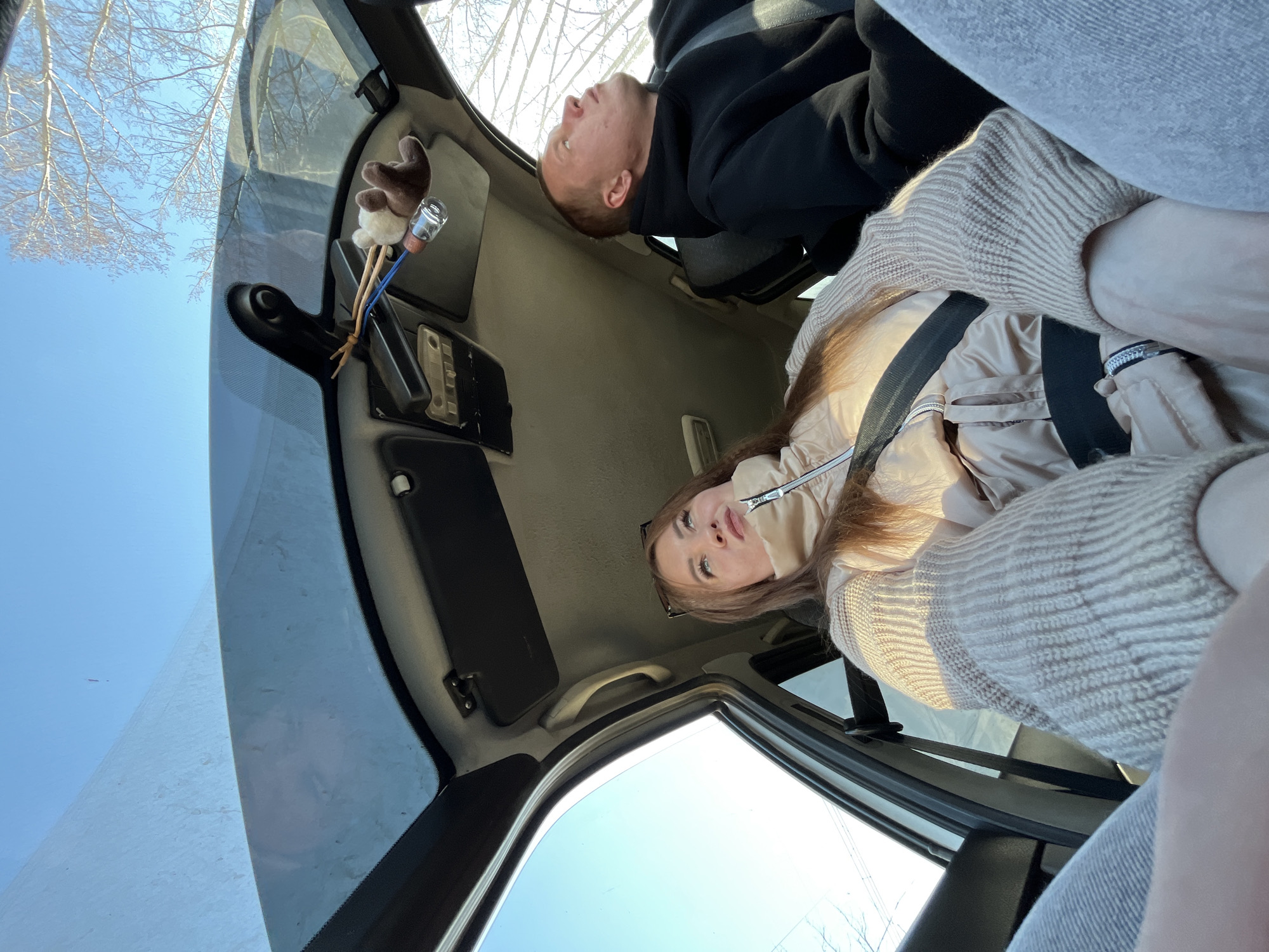 Two people are in a car, a woman facing the camera with a seatbelt on wearing a beige jacket and sweater, and a man with his face turned upwards wearing a black hoodie. The car is driving outdoors on a clear day. An air freshener dangles from the rearview mirror. Branches from bare trees are visible through the windshield. The location is Chaikovsky, Russia, based on the information provided.