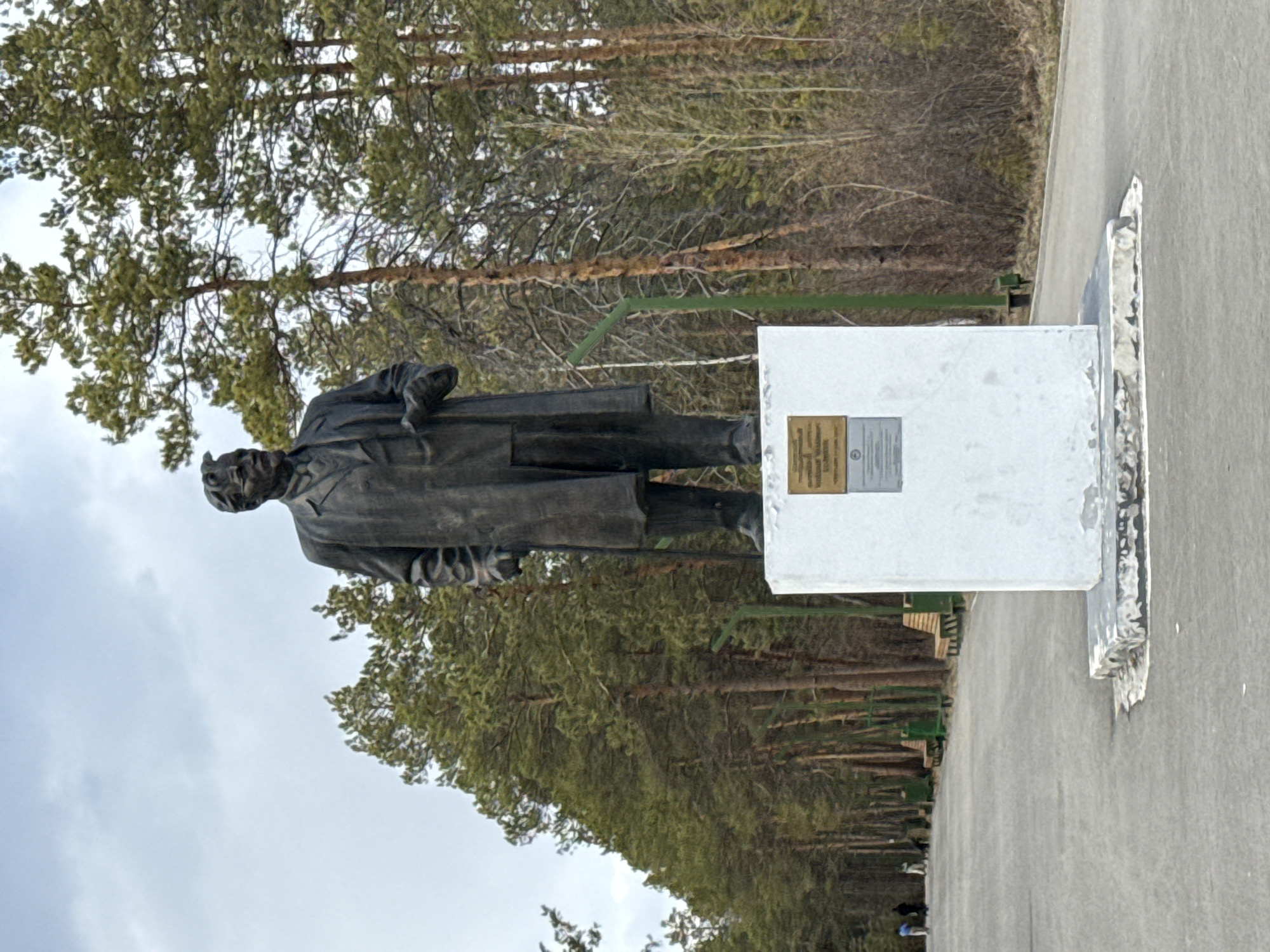 A statue of a man in a long coat stands on a white pedestal on a concrete pathway. The statue appears to be made of dark metal. The pedestal has a plaque on it. A forest of pine trees lines the background. The sky is overcast. The statue is in Yakutsk, Russia.