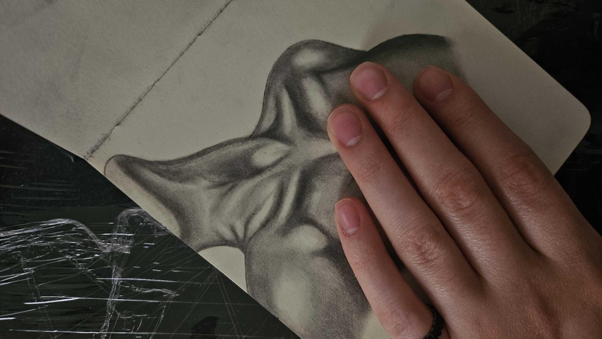 A close-up, overhead shot shows a graphite drawing of the upper torso and neck of a person, with a human hand positioned over the drawing. The drawing is on a light-colored paper, angled in the frame, with a dark, textured background that appears to be covered in crinkled plastic. The drawing depicts strong shading to create the illusion of muscle definition and bone structure, particularly around the clavicle, sternocleidomastoid muscles, and deltoids. The hand in the foreground has fair skin and is slightly turned, with the fingers extended across the lower portion of the drawing. The fingernails are clean and unpolished, with a faint white crescent visible at the base of each. A dark, textured ring is present on the ring finger. The lighting in the image is uneven, creating shadows and highlights on both the drawing and the hand. The scene appears to be indoors, likely in an art studio or workspace, given the nature of the subject matter and the presence of drawing materials. The overall impression is that of an artist's work in progress or a staged photograph showcasing the artwork.