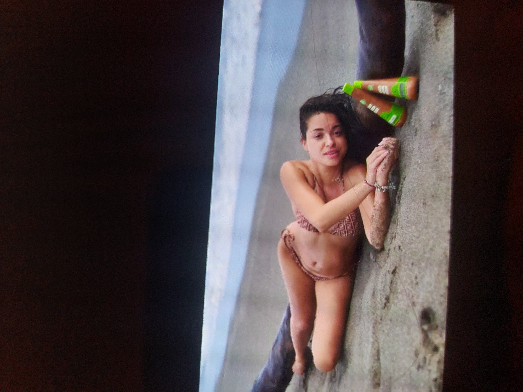 A young woman with dark, wavy hair is pictured kneeling on a rough, concrete or rocky surface, likely outdoors near water in Tsarevo, Bulgaria. She is wearing a patterned light brown or beige bikini. Her hands are clasped together in front of her, resting on the ground, with some sand or dirt visible on her hands and forearms. She looks directly at the camera with a soft smile. She wears a delicate necklace and several bracelets on her left wrist.
Behind her head, leaning against a dark, possibly wooden, vertical structure, are two brown bottles with bright green caps and labels. The brand "OGX" is visible on both bottles. To the left of the frame, a body of water, possibly the sea or a large pool, is visible. The scene appears to be set during the daytime under bright conditions. The overall image suggests a relaxed moment outdoors. The photograph itself appears to be an image of a screen, with dark borders on the left and right.