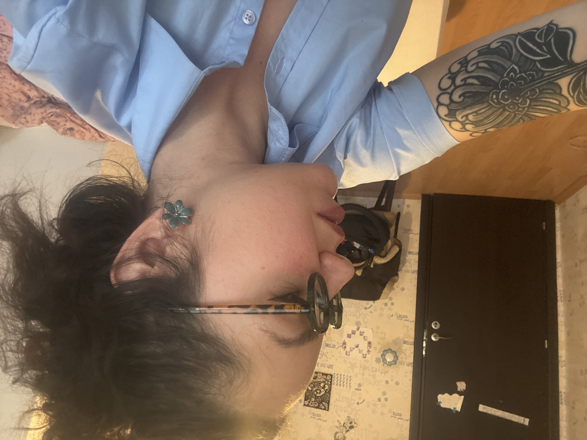 A woman with dark curly hair, wearing glasses and a light blue button-down shirt with an open collar, is visible in a rotated image. She has a floral earring in her left ear and a tattoo on her right arm. The background includes a door, patterned wallpaper, a chair with a jacket, and part of a bed. The location is unspecified, and Burgas, Bulgaria is not visibly relevant to the image.