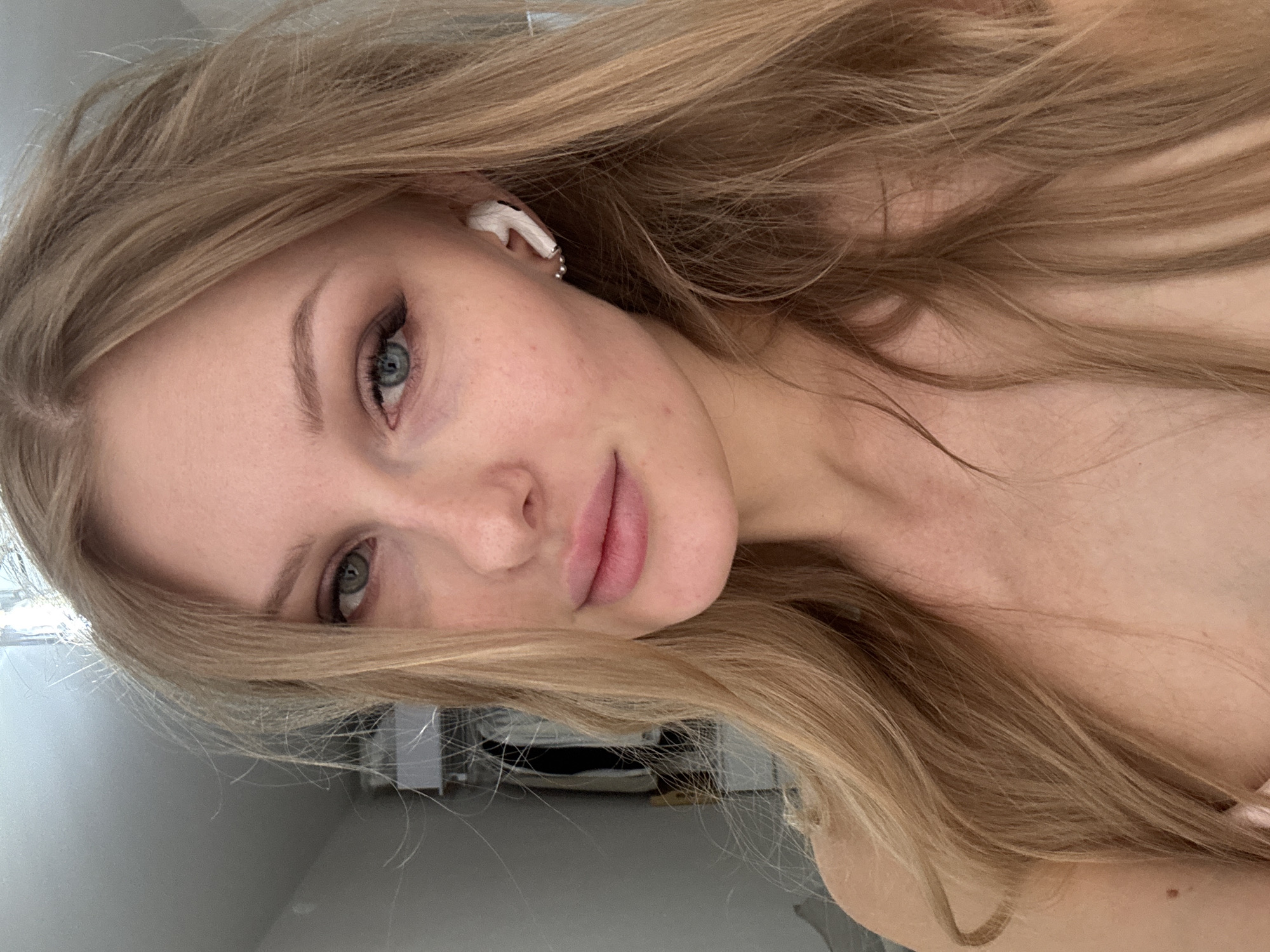 A blonde woman with light skin is centered in the frame, looking directly at the camera with a slight smile. She is wearing a white wireless earbud in her left ear and has light makeup on her eyes. Her long blonde hair falls around her face and shoulders. The background appears to be a sparsely decorated room, with a piece of furniture and a partially visible window or light source. The image was taken indoors with what appears to be a mobile phone camera. There is no indication that the photo was taken in Perm, Russia.