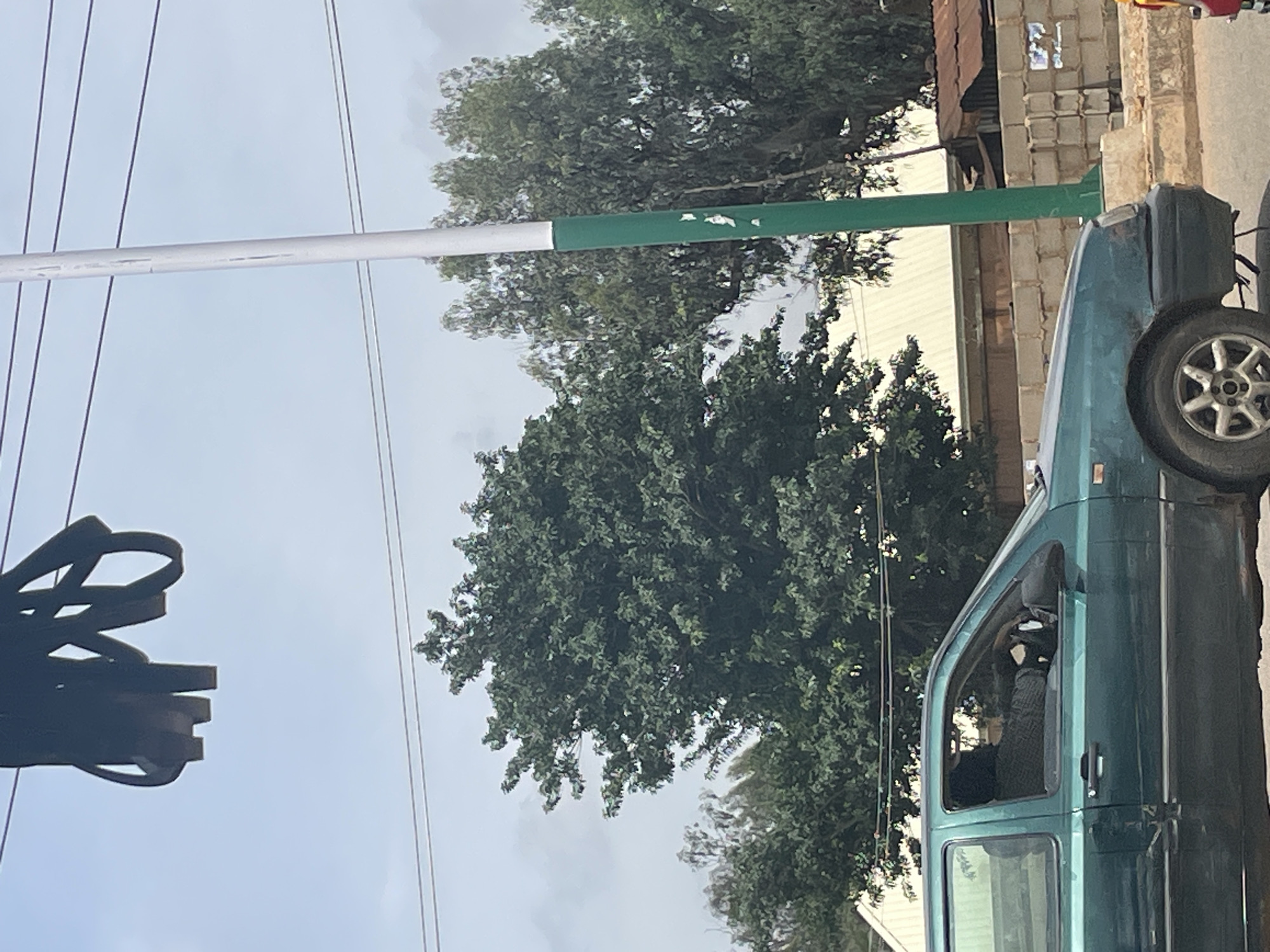 This is a slightly tilted, medium-shot outdoor photograph taken in Bukuru, Nigeria. The main subjects are a green vehicle and a horizontal pole with white and green sections, which appears to be a traffic barrier or part of street infrastructure. Trees with dense green foliage are visible in the background.
The vehicle is a dark green, possibly an older model car, with its side and part of the front visible. The driver's side window is partially open, and a person wearing a dark head covering and a patterned shirt is visible inside the car. The vehicle's wheel is also visible.
The pole runs horizontally across the middle of the frame, with the left portion being white and the right portion being green. It appears to be attached to a vertical pole on the left, which is out of focus and partially obscured by a dark, abstractly shaped object that might be a traffic light or another piece of street furniture. In the background, beyond the trees, there are buildings constructed with cinder blocks and a corrugated metal roof, suggesting an urban or semi-urban environment.
The sky is overcast with some lighter patches, indicating a cloudy day. The lighting is diffused, suggesting it could be morning or late afternoon. There is no explicit text visible in the image. The overall impression is a snapshot of a typical street scene in Nigeria, focusing on the vehicle and the surrounding environment.