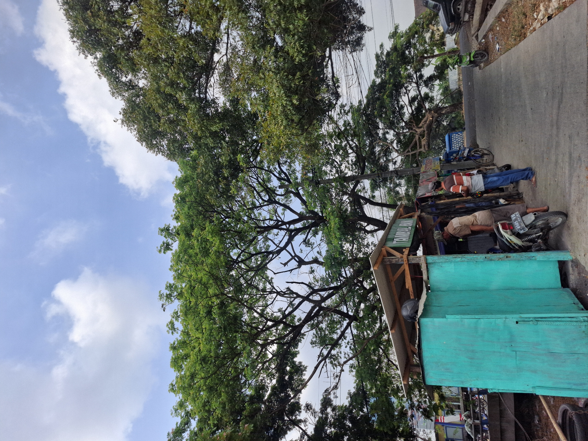 The image depicts a street scene in Sidoharjo, Indonesia, characterized by a clear sky with scattered clouds and lush green trees overhead. In the foreground, a small roadside stall with a turquoise wooden structure and a corrugated metal roof is visible. Two men are standing near the stall. One, wearing a brown shirt and a cap, is attending to a parked motorcycle. The other man, dressed in a horizontally striped shirt and blue jeans, appears to be interacting with the first man. A motorcycle is parked on the paved road next to the stall. Further down the road, a green motorcycle is parked, and a dark-colored vehicle is partially visible. The environment suggests a typical Indonesian street with a mix of commercial activity and natural elements.