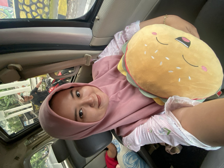 A woman wearing a pink hijab sits in a car, holding a plush hamburger pillow with a smiling face. She faces the camera and smiles. The car window is open, reflecting a man standing outside. She wears light pink pajamas with a lace trim. The car's interior includes gray seats and a seatbelt. The background shows glimpses of greenery and buildings through the car window.