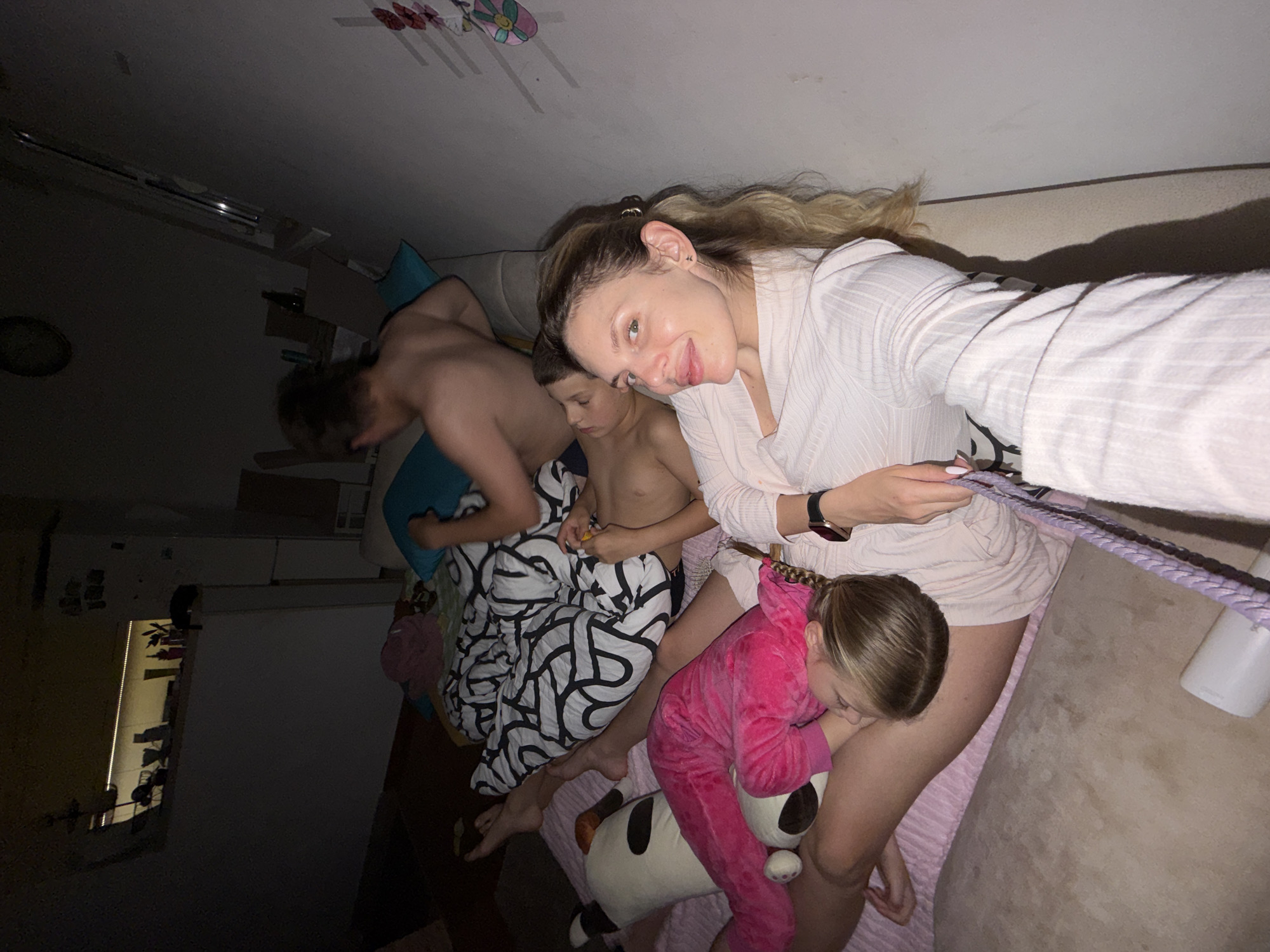 Four individuals are visible in the image. A woman in a light-colored robe holds up a cell phone to take a selfie. Next to her, a child in a pink jumpsuit embraces a plush panda toy. Two other individuals, both shirtless, lie under a patterned blanket. The environment appears to be an interior, likely a residential bedroom. No identifiable locations in Budva, Montenegro, are discernible from the visual data provided.
