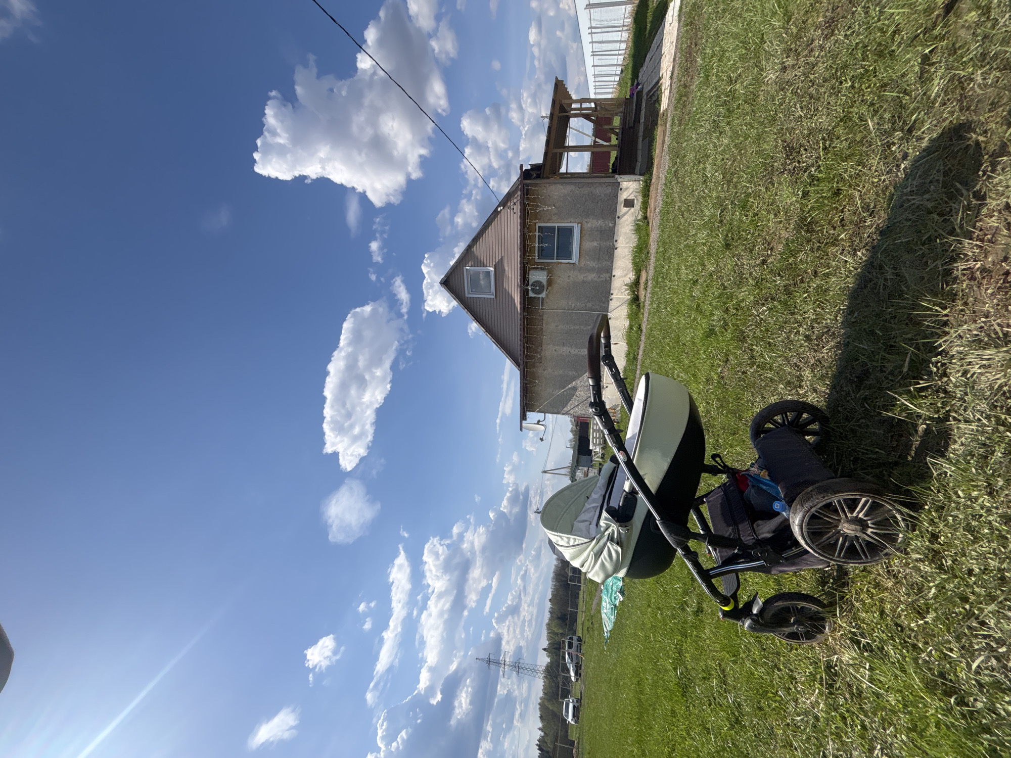 A color, full shot shows a black stroller in the grass in front of a one-story building on a bright, sunny day in Раздолье, Russia.
The stroller is black with an eggshell-colored bassinet. The wheels are dirty. Behind the stroller is a one-story building with pale green siding and a brown peaked roof. There is an air conditioning unit attached to the exterior wall near a square window. A glass greenhouse is attached to the back of the building. Powerlines run above the building. The grass is a bright green. The sky is blue with puffy white clouds. There is a field in the distance.