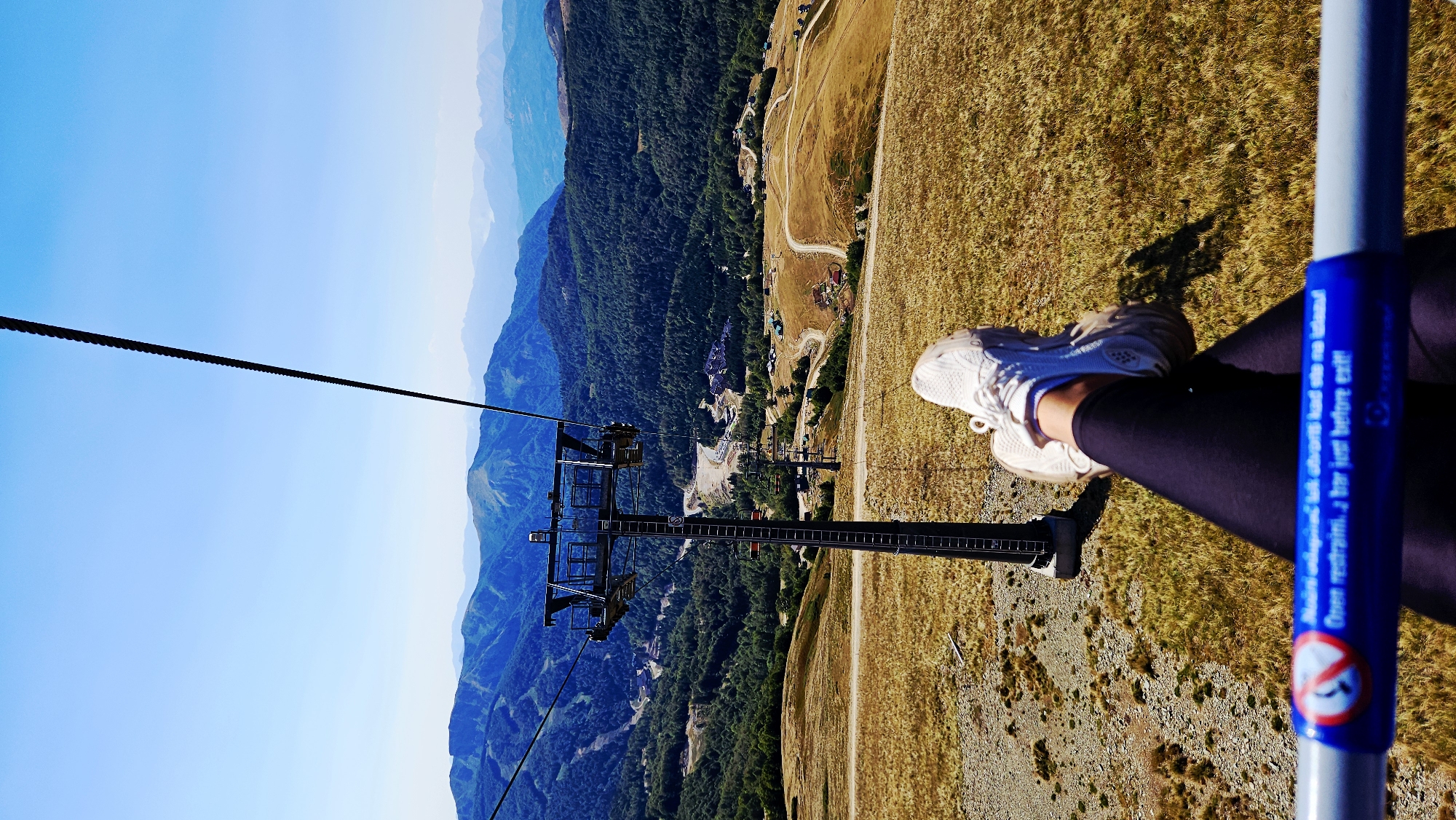 The image is taken from the perspective of someone riding a ski lift on a sunny day. In the foreground, the legs of a person wearing white sneakers and dark leggings are visible, resting on the ski lift bar. The ski lift bar has a blue and white warning sticker with text in both an unknown language and English: "Open restraints a bar just before exit!".
In the mid-ground, a tall ski lift tower with a complex structure at its top is prominent. Several other ski lift towers and cables extend into the distance. The landscape consists of dry, golden-brown grass on the slopes, with patches of rocky terrain. Further into the background, a lush green forest covers the hillsides, leading up to distant, hazy blue mountains under a clear blue sky.
The location is identified as Sedlačka gora, Montenegro. The scene depicts a scenic mountain environment, likely during the summer or early autumn, as indicated by the dry grass and clear weather. The ski lift infrastructure suggests that this location is a ski resort, currently not in operation for winter sports.