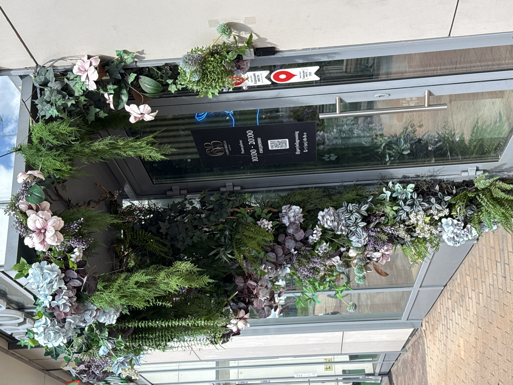 The image shows the entrance to a business called "Garry & Bella Grooming" in Moscow, Russia. The entrance is decorated with an abundance of artificial greenery and flowers, creating a lush, arching canopy over the doorway. The doorway itself is a glass door with a silver handle. On the glass door, there is a black sign with white text and a QR code. The sign displays the business name, operating hours (10:00 - 20:00 daily), social media handle (@garrybellagrooming), and a phone number. There are also two red and white stickers on the wall next to the door, one with a red location pin icon and text that appears to be in Russian. The sidewalk in front of the entrance is paved with brick. The building facade is light-colored. The sky is visible above the entrance, and it is a bright, sunny day.