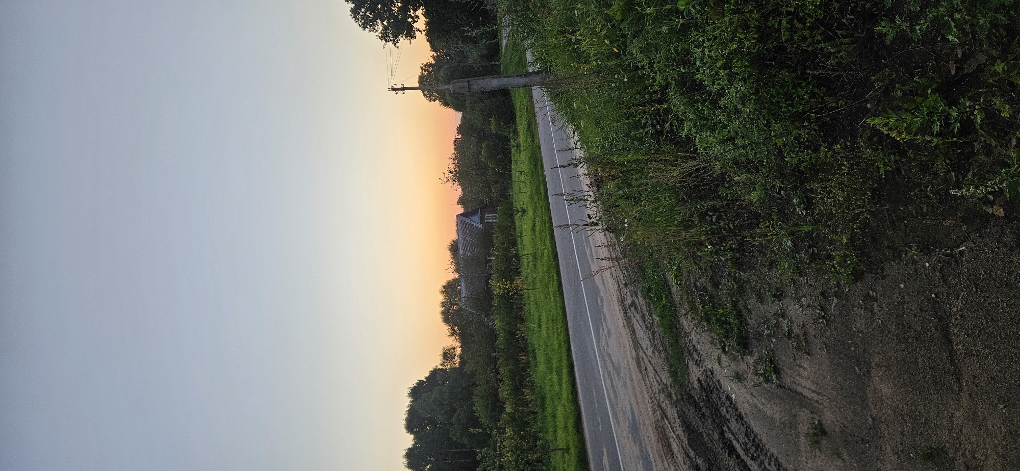 A vertical, medium-shot photograph captures a rural road in Mazalawski Sel'ski Savet, Belarus, at dusk. The sky transitions from a pale blue at the top to a warm orange and pink hue near the horizon, indicating the setting sun. A paved road with a white dividing line cuts diagonally from the lower left corner towards the center of the frame. The road's shoulder is composed of dirt and mud, with visible tire tracks on the left side. Lush green grass and vegetation line the opposite side of the road. In the middle ground, a cluster of trees and bushes partially obscures a rustic, dark-colored house with a gray roof. A solitary concrete utility pole stands on the right side of the road, with wires extending from it. The foreground is dominated by overgrown green plants and dry grass. The overall scene depicts a tranquil, rural landscape under a twilight sky.