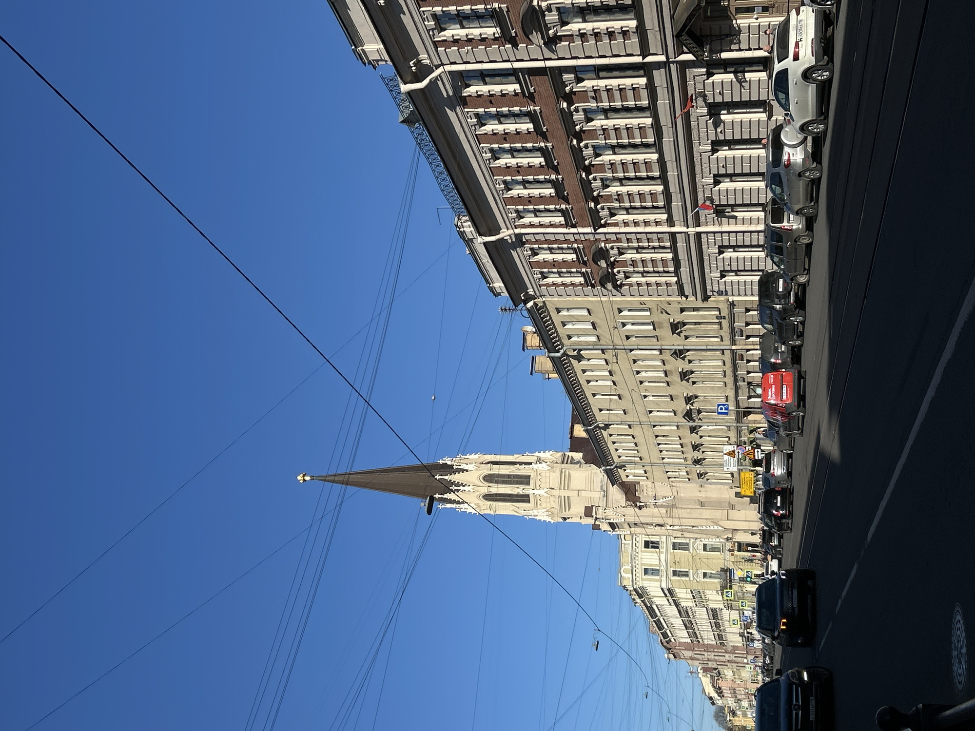 The image captures a vibrant street scene in Saint Petersburg, Russia, under a clear, bright blue sky, suggesting a sunny day, likely midday or early afternoon. The perspective looks down a wide street flanked by substantial buildings.
On the left side of the mid-ground, a prominent and ornate building, likely a church or cathedral, stands tall with a slender, dark-tipped spire reaching towards the sky. Its facade is light-colored and features Gothic-style architectural details, including pointed arches, decorative windows, and intricate stonework.
Along the right side of the street, a continuous row of multi-story buildings lines the road. These structures exhibit classical European architecture with numerous windows, cornices, and repeating decorative elements. Overhead, a dense network of black wires, likely for trams or trolleybuses, stretches across the sky.
The street itself is active with vehicles. Several cars, including sedans and SUVs of various colors (white, silver, black), are parked bumper-to-bumper along the right curb. Other cars are seen driving in the foreground and mid-ground. A red truck, possibly a municipal vehicle, is visible further down the street, with white Cyrillic text that appears to include "АВТО". Among the buildings, a blue street sign with a white 'P' indicates parking. Visible license plates on cars, such as one with the "178" region code, confirm the location as Saint Petersburg. The scene depicts a typical, bustling urban environment.