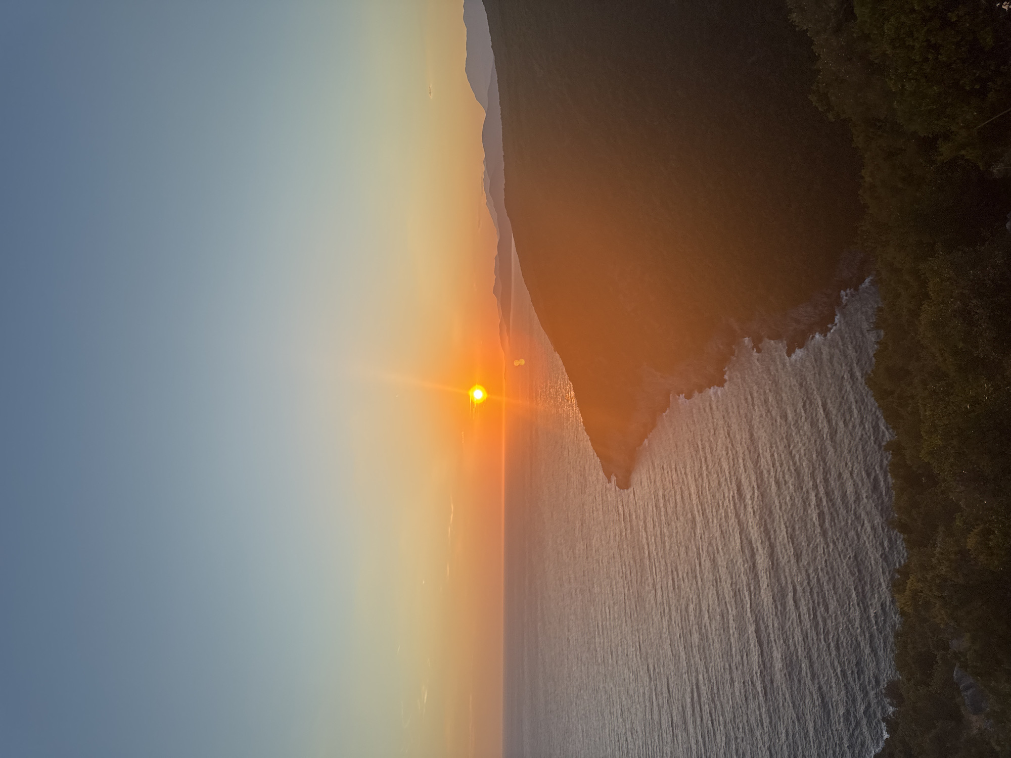 The image depicts a scenic sunset over the Adriatic Sea, with the coast of Zagora, Montenegro visible in the foreground. The sun, a bright orange orb, is positioned near the horizon, casting a warm glow across the sky and water. The sea is calm, with gentle waves reflecting the golden hues of the sunset. A lush, green coastline forms a prominent feature of the landscape, with a winding path leading down towards the sea. In the distance, a range of mountains can be seen silhouetted against the vibrant sky. The overall atmosphere is one of tranquility and natural beauty. There are no people or animals visible in the image.