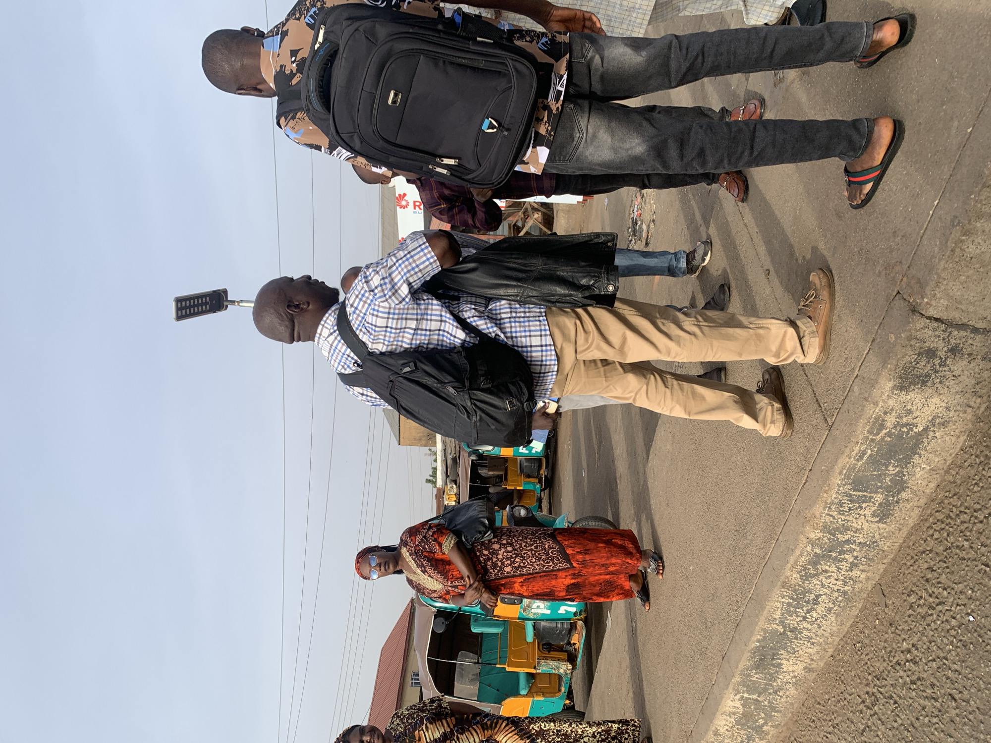 The image captures a street scene in Jos, Nigeria. In the foreground, a woman wearing an orange and brown patterned dress and blue sunglasses stands with her arms crossed. Beside her and partially obscured is another woman in a similar patterned dress. Behind them are several green and yellow auto-rickshaws, also known as "Keke Napeps". Moving towards the back of the image are two men with backpacks. The first is wearing a blue plaid shirt and khaki pants, carrying a black jacket over his shoulder. The second is wearing a patterned shirt and dark jeans. Overhead, power lines crisscross the sky. A sign with a red logo is visible in the midground. The scene is on a paved street with a concrete curb.