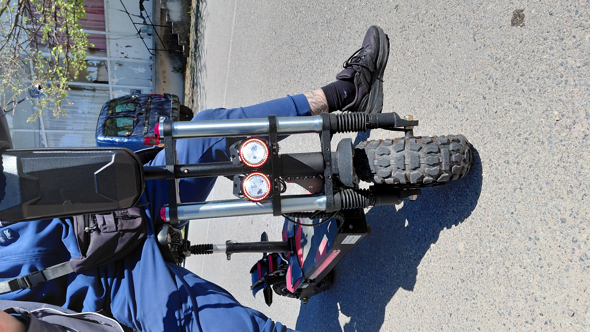 The image shows a person sitting on an electric scooter on an asphalt road in an urban environment. The scooter features two large front lights, a luggage rack, and a heavy-duty tire. The person is wearing a blue jacket, dark-colored pants, and black shoes with socks. A blue car is visible in the background near a building with peeling paint. A tree with green leaves is also visible in the upper left corner of the frame.