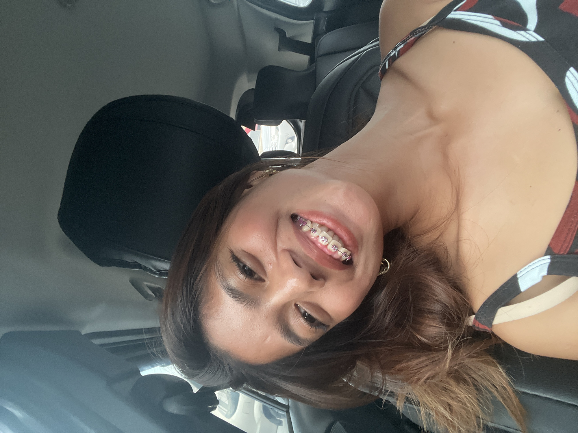 A young woman with brown hair and braces smiles widely while sitting in the passenger seat of a car. The car appears to be parked in a lot, as indicated by other vehicles visible through the windows. The woman is wearing a tank top with a geometric pattern. Her left earring is a small gold hoop, and there's a hint of what looks like glitter on her upper lip. The interior of the car is dark, with black leather seats and headrests. The overall scene suggests a casual moment captured during a day out.