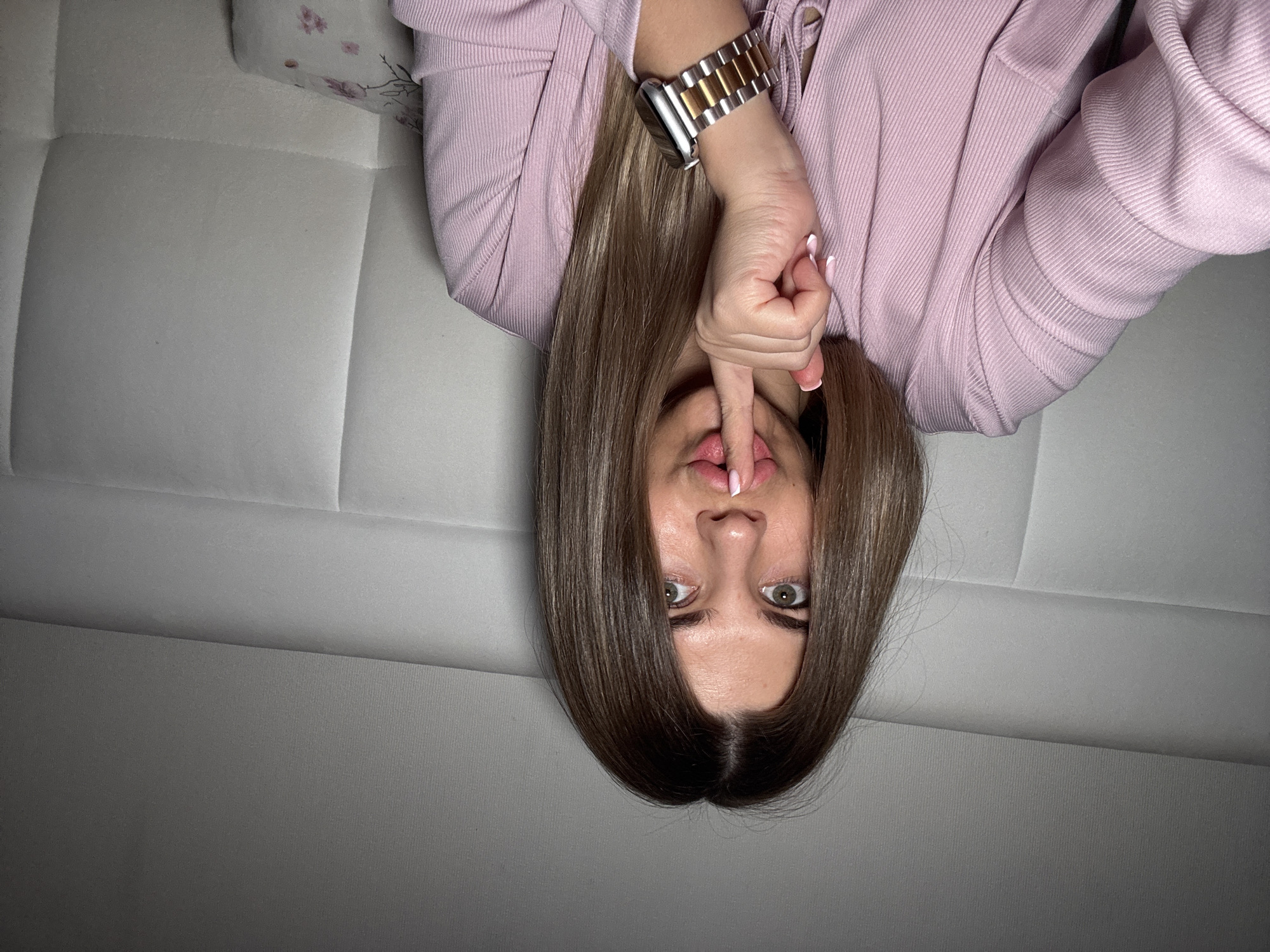 A woman with long, straight, light brown hair is lying upside down on a white upholstered couch. She is wearing a pink, long-sleeved, ribbed shirt and a stainless steel wristwatch with a gold band on her left wrist. Her left index finger is placed vertically against her lips in a "shush" gesture. Her eyes are open and looking directly at the camera. The background is a gradient of light to dark gray. This image may be from Chaikovsky, Russia.