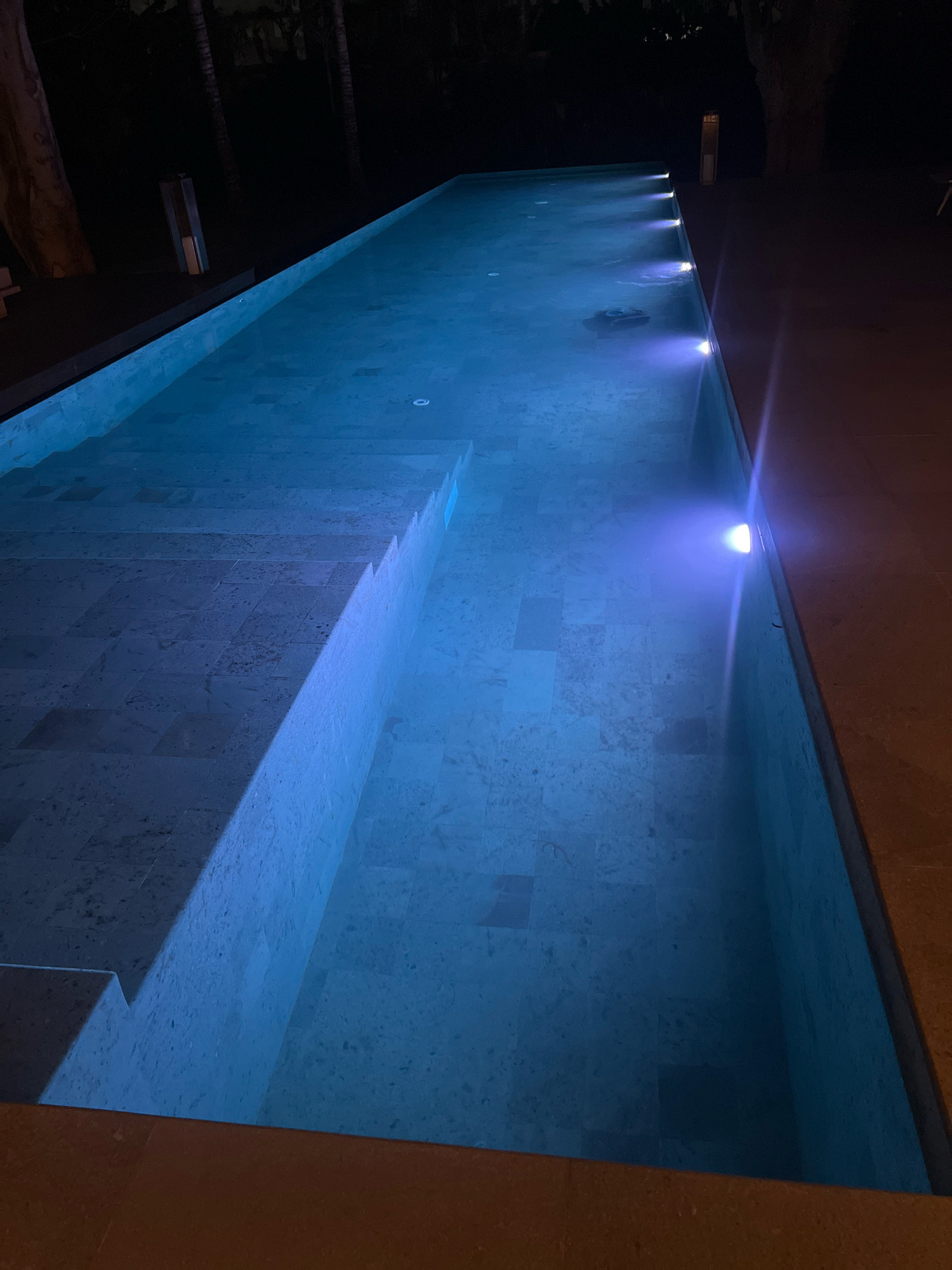The image depicts a long, rectangular swimming pool at night. The pool is illuminated by blue underwater lights, creating a cool glow. The water appears clear, and the pool floor is tiled with light-colored stone. A set of wide, shallow steps descends into the pool on the left side. The pool is surrounded by a dark tiled patio area. In the background, silhouetted palm trees and dark foliage are visible against a night sky. The overall atmosphere is tranquil and modern. This scene is located at The Layar, a luxury villa resort in Ngaglik, Indonesia.